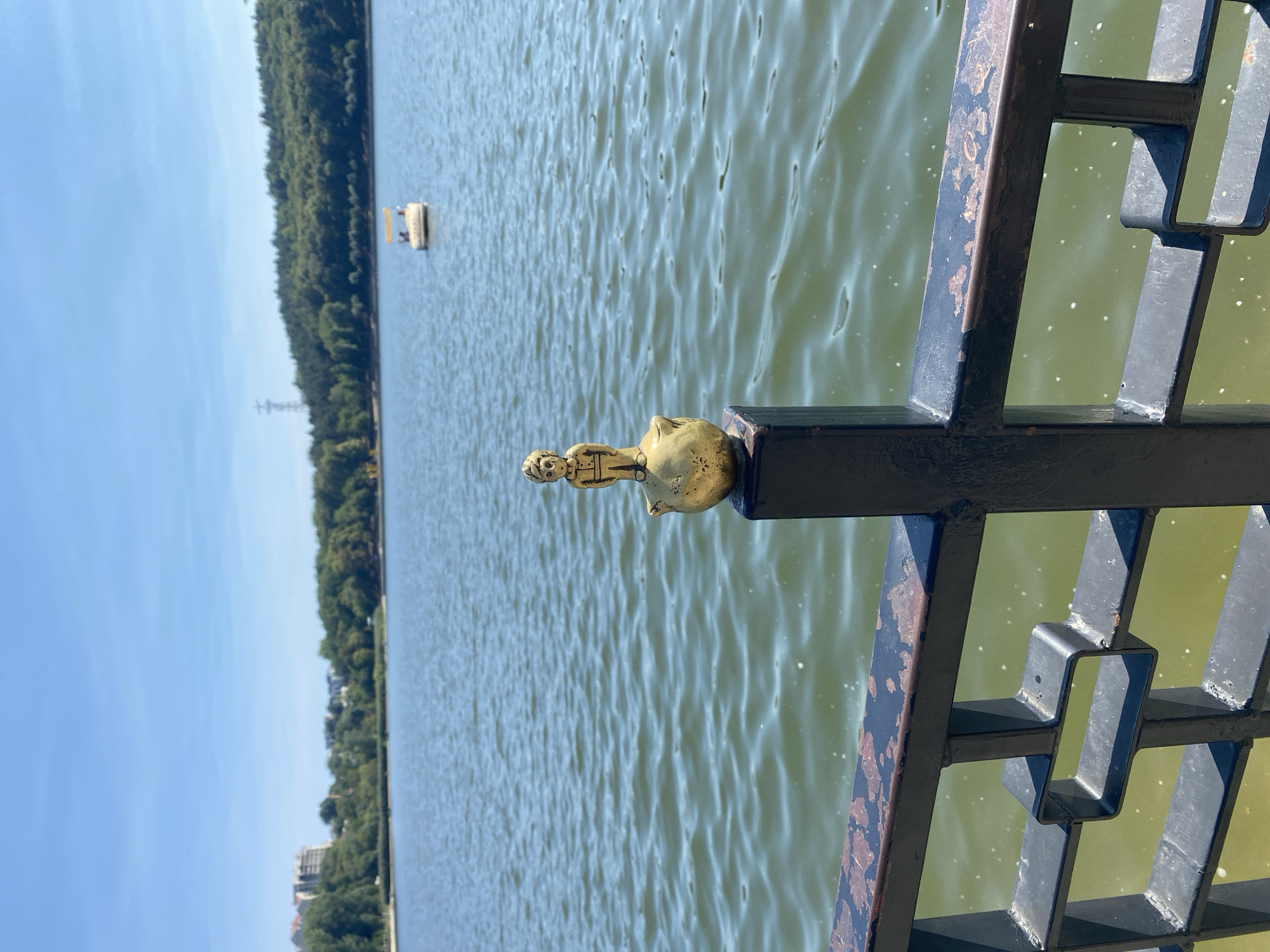 The image shows a small, golden statue of a boy standing on a sphere, perched atop a black metal fence post. The fence is in the foreground, with a dark railing and patterned infill. Behind the fence, a wide body of water stretches into the distance. The water is choppy, with small white flecks visible on its surface, possibly reflections or spray. In the middle distance, a small white boat with a yellow canopy is visible on the water. The background consists of a tree-lined shore, with a distant hill covered in dense green forest. Buildings are visible on the far left of the shore. The sky is a clear, bright blue. The overall scene suggests a sunny day by a lake or large body of water. While the exact location in Chisinau, Moldova cannot be definitively determined from the image alone, the presence of a lake, park-like surroundings, and potential urban development in the background are consistent with areas like the Valea Morilor Park, which features a lake and is a popular recreational spot in the city.