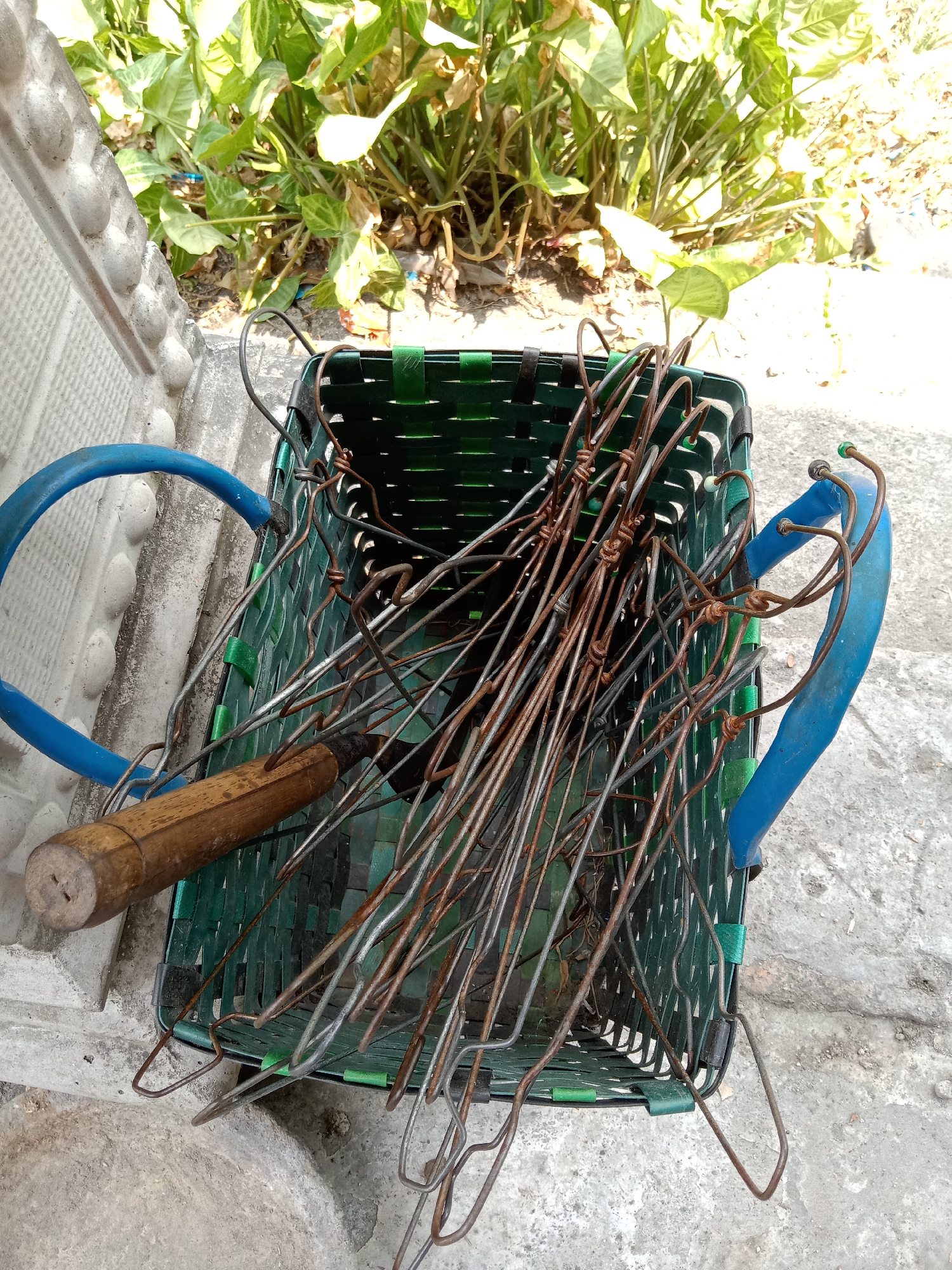 The image shows a green woven basket filled with rusty metal skewers and a wooden-handled tool, possibly a grill brush or scraper. The basket has two blue handles, one of which is partially visible on the left side of the frame, and the other on the right. The basket and its contents are resting on a concrete surface, with some greenery and foliage visible in the background. The scene appears to be outdoors, possibly in a yard or garden, with the sunlight casting shadows. The overall impression is one of an object stored outdoors, perhaps awaiting use or after use. The location in Jumput, Indonesia is not discernible from the visual information in the image.