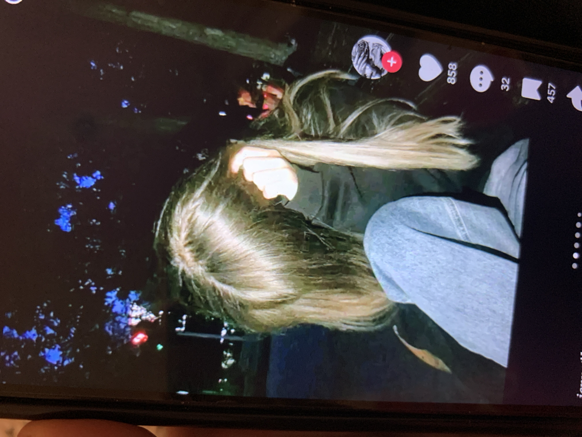 The image, captured on a mobile device, displays a TikTok video interface. The video features a person with long blonde hair, partially obscured by their hand, wearing a dark hoodie and light blue jeans. They are seated, with their left leg bent and covered by a denim garment. The background is dark, with blurry lights suggesting an outdoor, nighttime setting. Notably, the video interface elements, including a profile icon with a red plus sign, a heart icon with "858" likes, a speech bubble icon with "32" comments, and a bookmark icon with "457" shares, are visible on the right side of the screen. The image does not provide any discernible information about the city of Lymanka, Ukraine, or any specific location details within it.