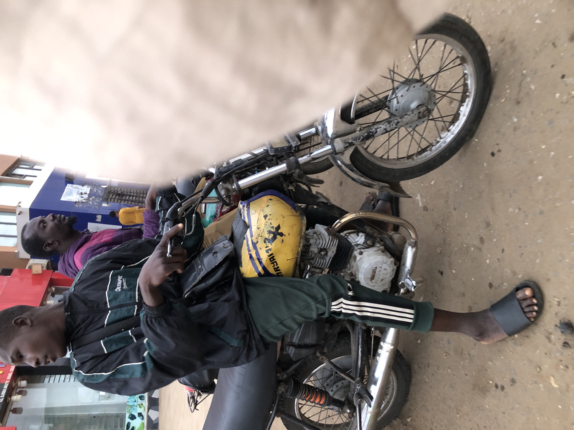 In the city of Ojodu, Nigeria, a young Black man is seated on a motorcycle, his right hand resting on the handlebars. He wears a black jacket with green and white accents, dark green track pants with white stripes, and black slide sandals. The motorcycle has a yellow fuel tank with blue stripes and text that appears to say "OKAYAY". A second Black man, wearing a purple hooded jacket, is seated behind the driver, and a third person in a yellow shirt is visible in the background. The environment appears to be an urban street with buildings and a dusty road.