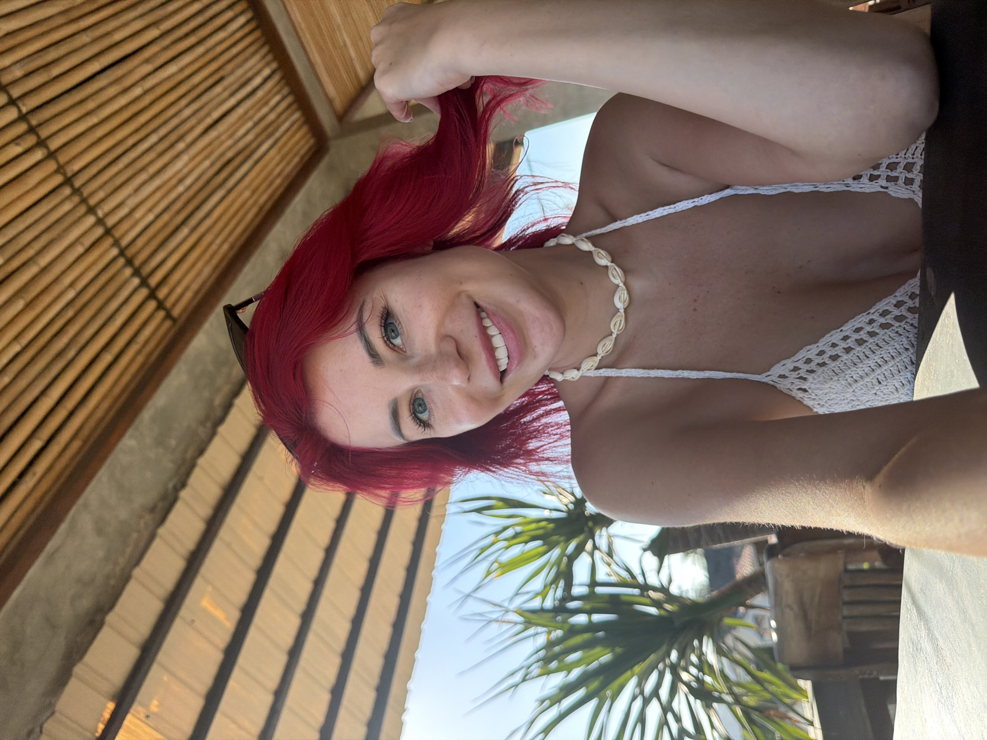In Kuta, Indonesia, a woman with bright red hair and blue eyes is captured in a selfie. She is wearing a white crocheted bikini top and a seashell necklace. She is sitting at a table under a thatched roof structure, with palm trees and a clear blue sky visible in the background. She is smiling and holding a strand of her hair.