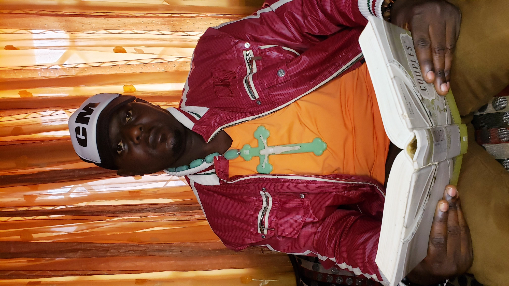 In Sabon Tasha, Nigeria, a man is seated, holding an open book. He is wearing a red jacket with white and green trim, an orange shirt, and a white baseball cap with "CM" printed in black. A large, light green cross pendant hangs from a beaded necklace around his neck. The background consists of orange curtains with a patterned design. The man's hands are holding the book, which appears to be a devotional titled "Couples." He is looking at the book.