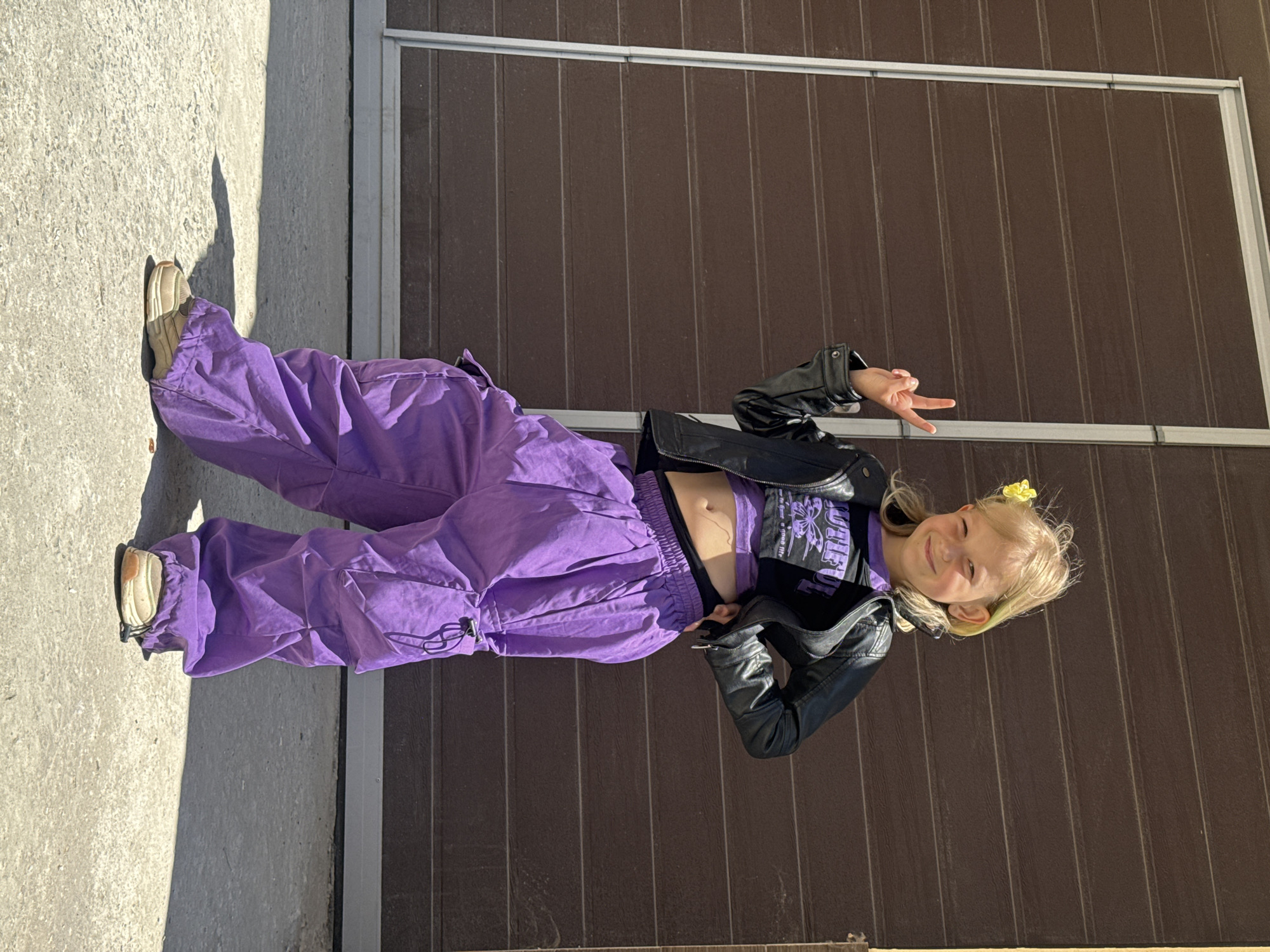 A young Caucasian girl with blonde hair stands in front of a brown, vertically-paneled wall. The setting appears to be outdoors on a sunny day, based on the bright lighting and shadows on the adjacent concrete surface.
The girl is wearing purple pants, a black t-shirt with a purple design, and a black leather jacket. She is also wearing light-colored sneakers and has a yellow flower in her hair. She is making a peace sign with her fingers and smiling at the camera.