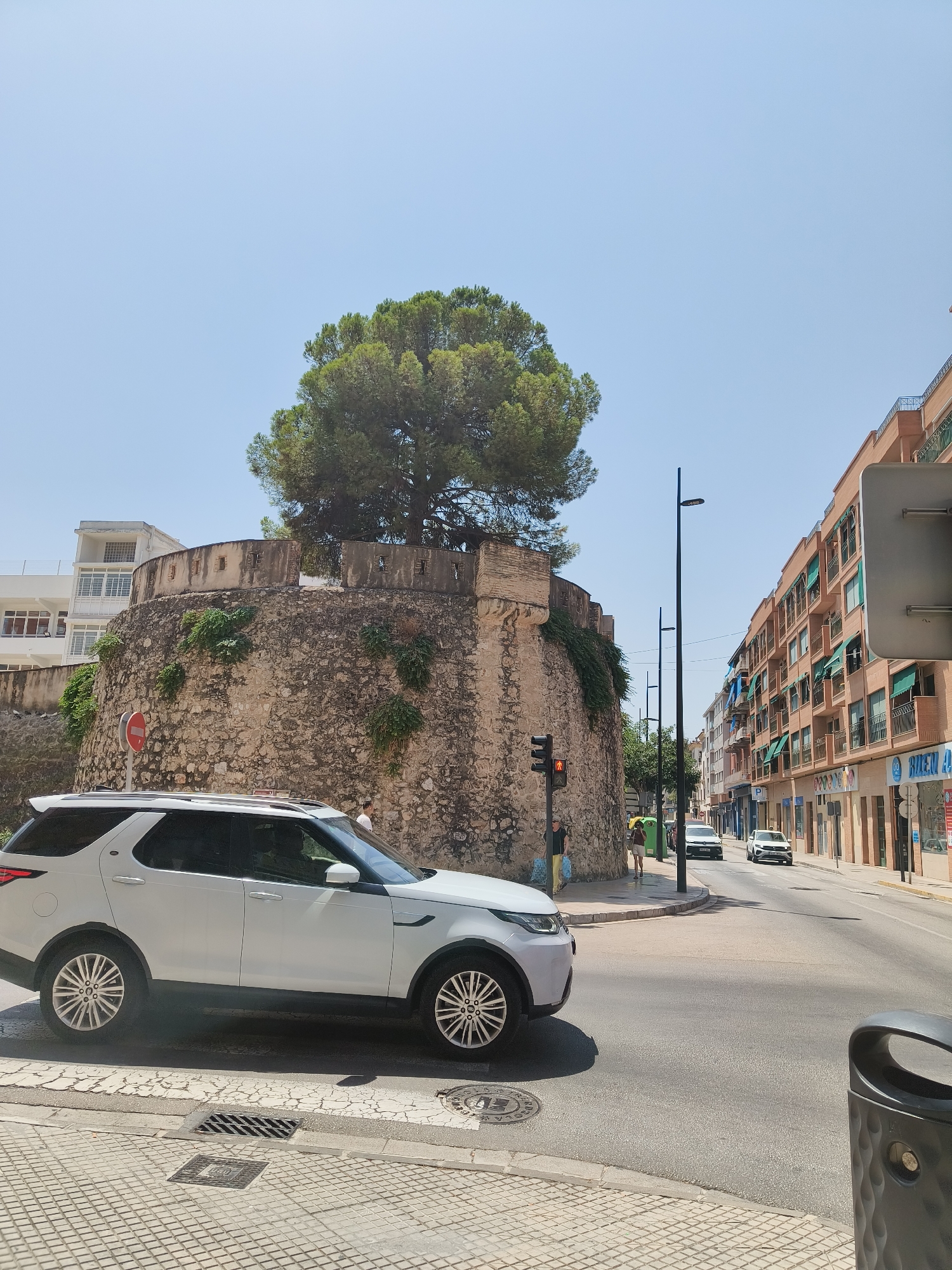 The image depicts a street scene in Gandia, Spain, focusing on a section of an old stone fortification topped with a mature tree. In the foreground, a white Land Rover Discovery is parked or stopped at a street intersection. The fortification, made of rough-hewn stones, features several small openings along its upper edge. Greenery grows on the side of the fortification.
To the right of the fortification, a street runs into the distance, lined with multi-story buildings featuring balconies with green awnings. Several cars are visible on the street, including a silver Volkswagen Golf and a white car further down. Streetlights and traffic signals are also present. A pedestrian crossing with a red pedestrian signal is visible near the fortification.
The sky is clear and blue, indicating a sunny day. The overall environment suggests an urban setting with historical elements.