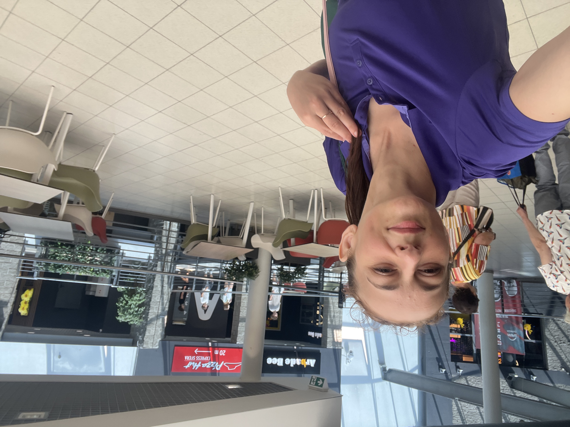 A woman, wearing a purple polo shirt, takes a selfie in a shopping mall. In the background, another woman in a colorful striped shirt walks away, and a man in grey pants and a white shirt walks past her. Several tables and chairs are visible in the seating area of a food court, with a Pizza Hut Express and Arcade Bee store in the distance. The floor is tiled in a light color.