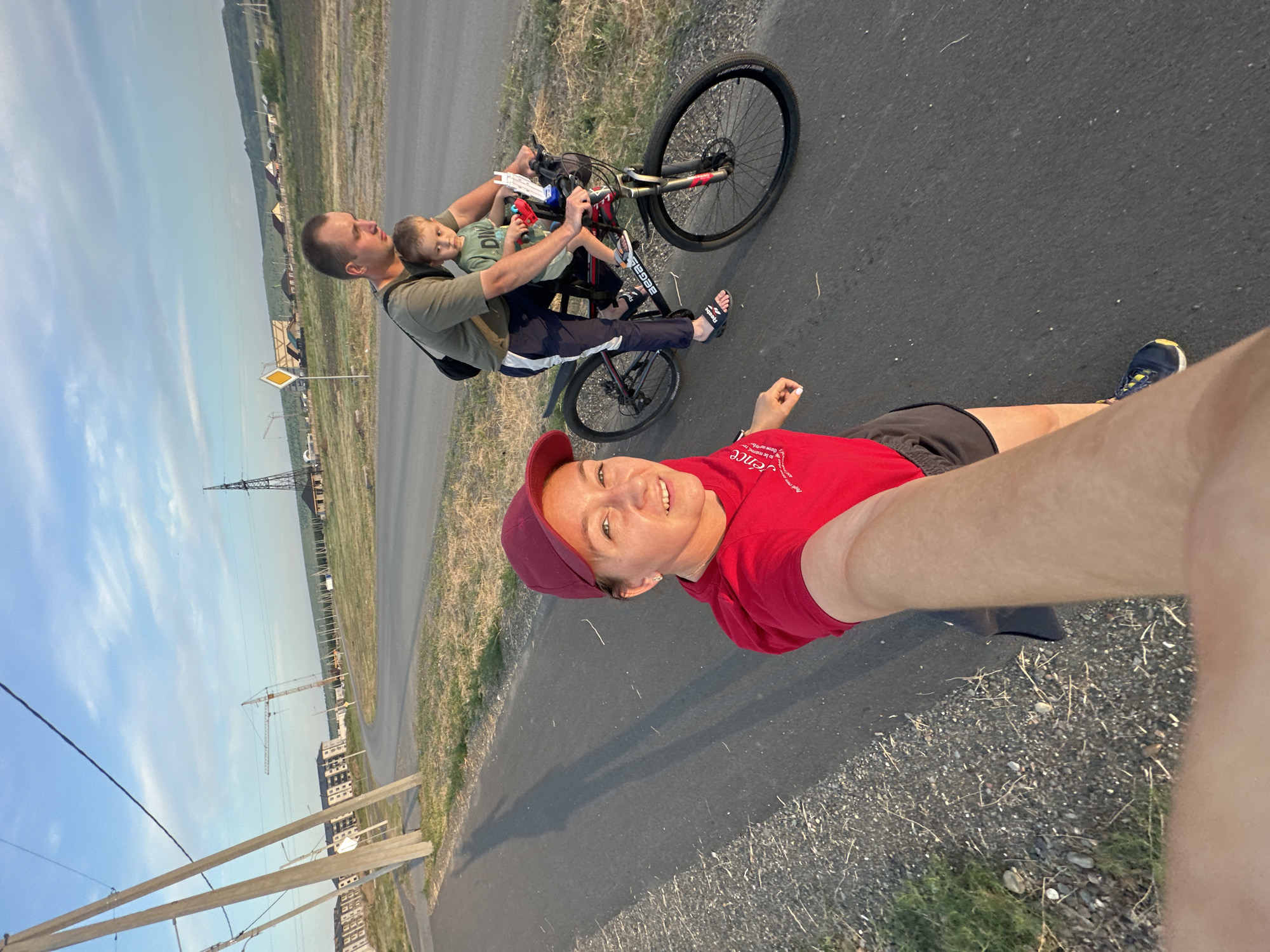 A selfie-style video frame shows a woman in the foreground on a paved path. She is wearing a red hooded shirt, dark shorts, and a red baseball cap. Her left arm is extended towards the camera, and her right hand is visible. In the background, a man is riding a bicycle with a young child seated in a child seat on the front. The man wears a dark t-shirt and dark pants with white stripes. The child wears a light green t-shirt. The bicycle is black and red. The background shows a paved road, a grassy shoulder, utility poles, and distant buildings and a construction crane under a blue sky with some clouds. The image appears to be taken during the golden hour, with long shadows cast on the path. This location is identified as Bely Yar, Russia.
