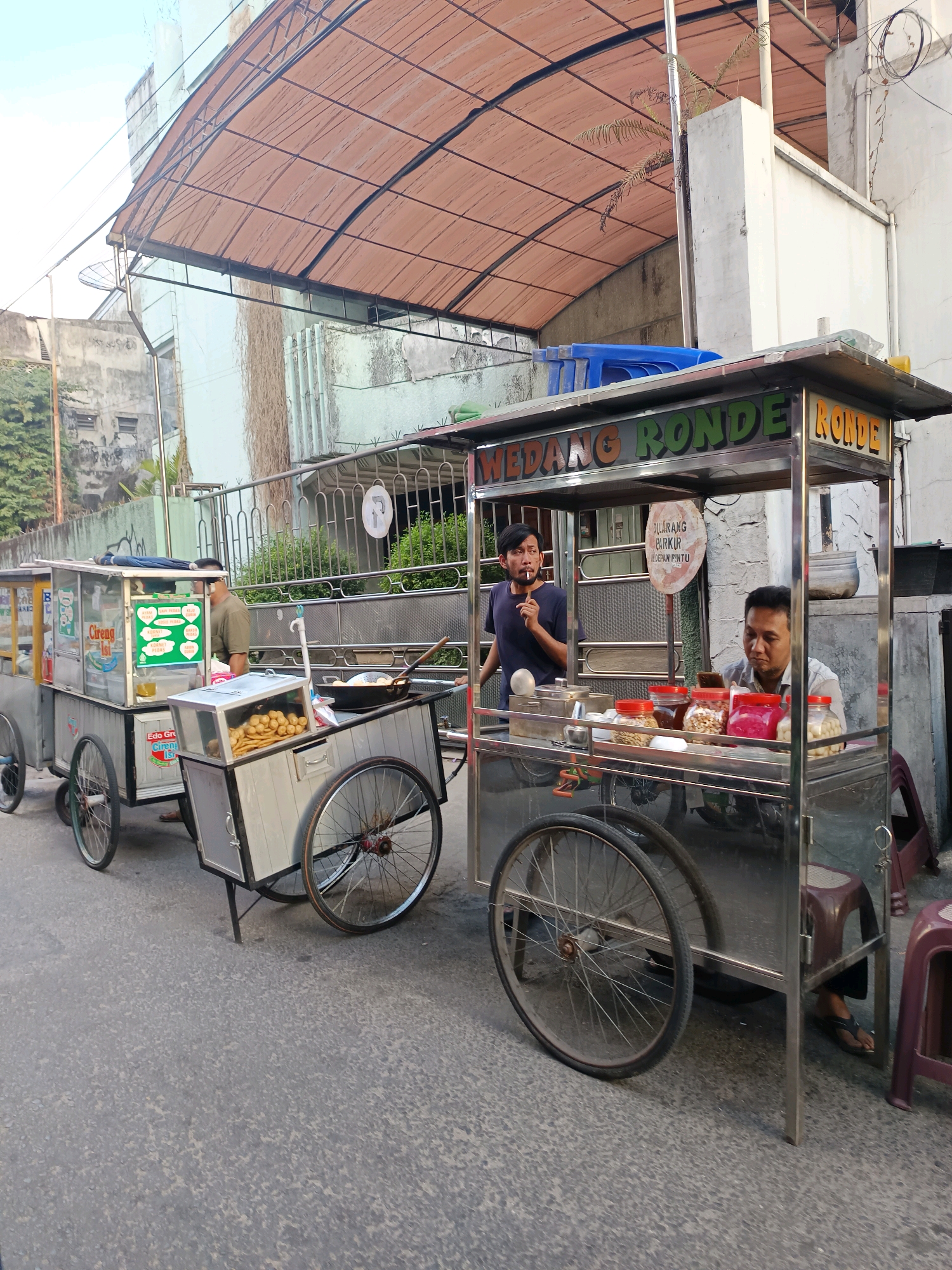 In the city of Yogyakarta, Indonesia, two food carts are positioned on a paved street. The cart on the right is larger and made of stainless steel, featuring a sign that reads "WEDANG RONDE RONDE". A man with dark hair and a black t-shirt is standing behind the cart, holding a cigarette. To his left, another man with a light-colored shirt is seated, looking down at something in his hands. The cart displays an assortment of jars containing various food items. In front of the cart, two red plastic stools are visible.
To the left of the main cart is a smaller food cart with a light-colored body and transparent glass panels. This cart has a sign that appears to advertise "CIRENG ISI" and lists several menu items. A man wearing a green shirt and a light-colored mask is visible behind this cart. A deep frying pan containing food is placed on top of this cart. The street is made of asphalt, and a building with a light-colored facade and a reddish-brown awning covers the carts. Green foliage is visible behind the food carts. The scene appears to be set outdoors during the daytime.
