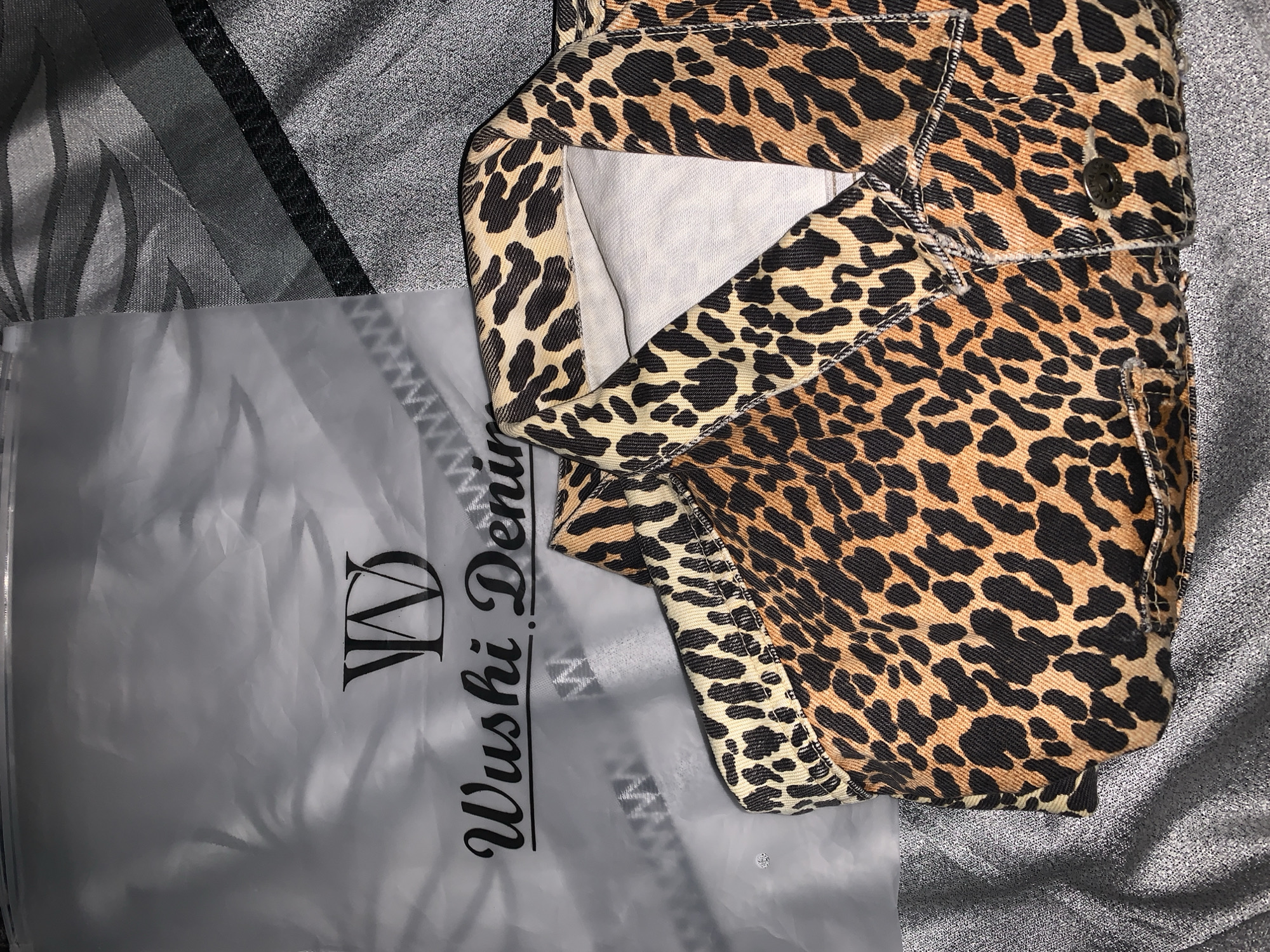The image shows a leopard print denim jacket lying on a silver, textured surface. Next to the jacket is a translucent plastic bag printed with "Wushi Denim" in black script. There is no visible indication of location, person, actions, or interactions. It is not possible to determine if the image was taken in Rafin Par, Nigeria.