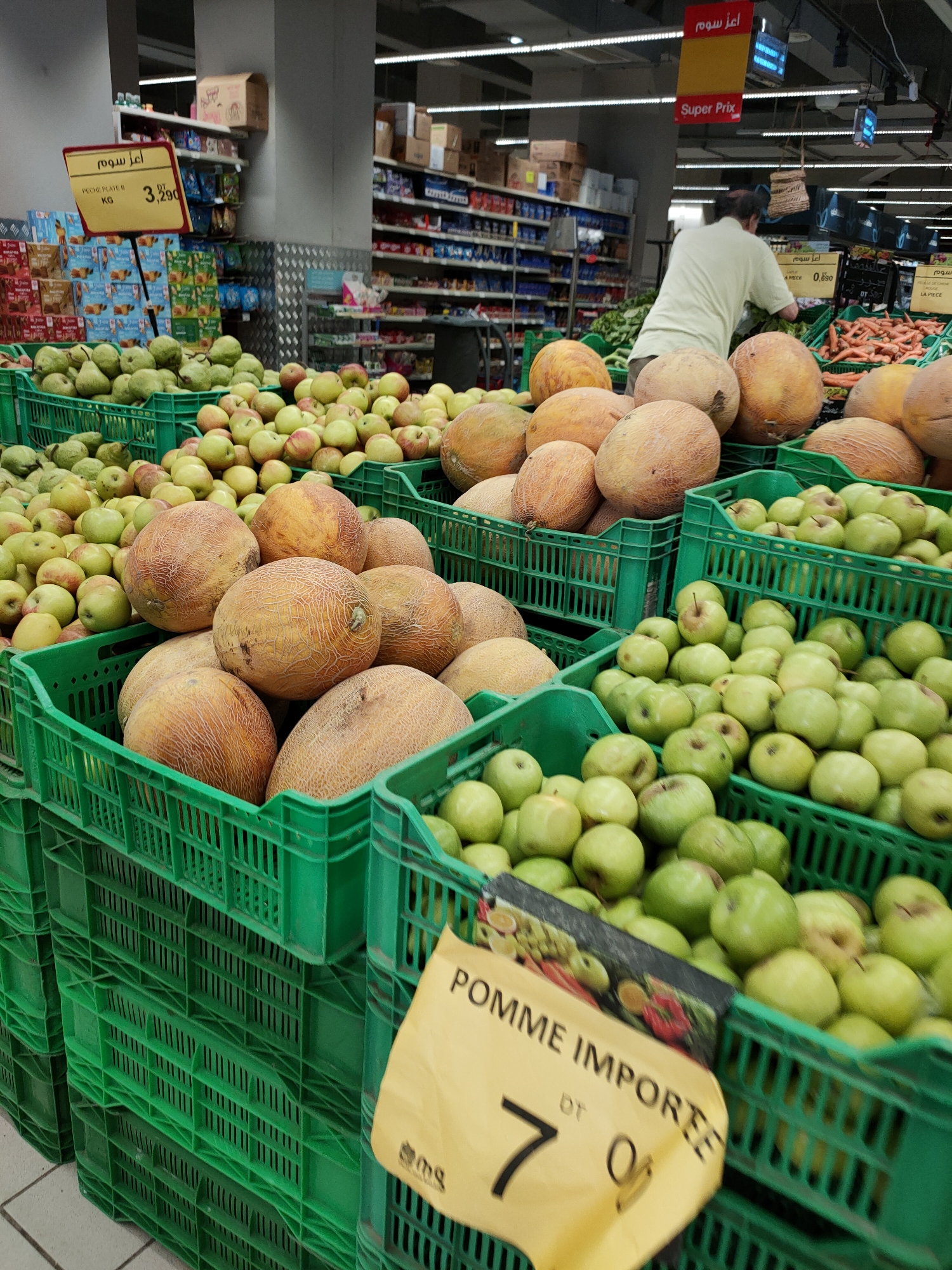 The image shows a produce display in a supermarket in Tunisia. In the foreground, green plastic crates are filled with various fruits, including melons, apples, and pears. A yellow price tag is visible in the lower right corner, displaying "POMME IMPORTEE" and a price of "7 DT". In the background, shelves are stocked with packaged goods. A man in a light-colored shirt is standing behind the produce, appearing to sort or arrange items. The overall environment suggests a retail setting with a focus on fresh produce.
