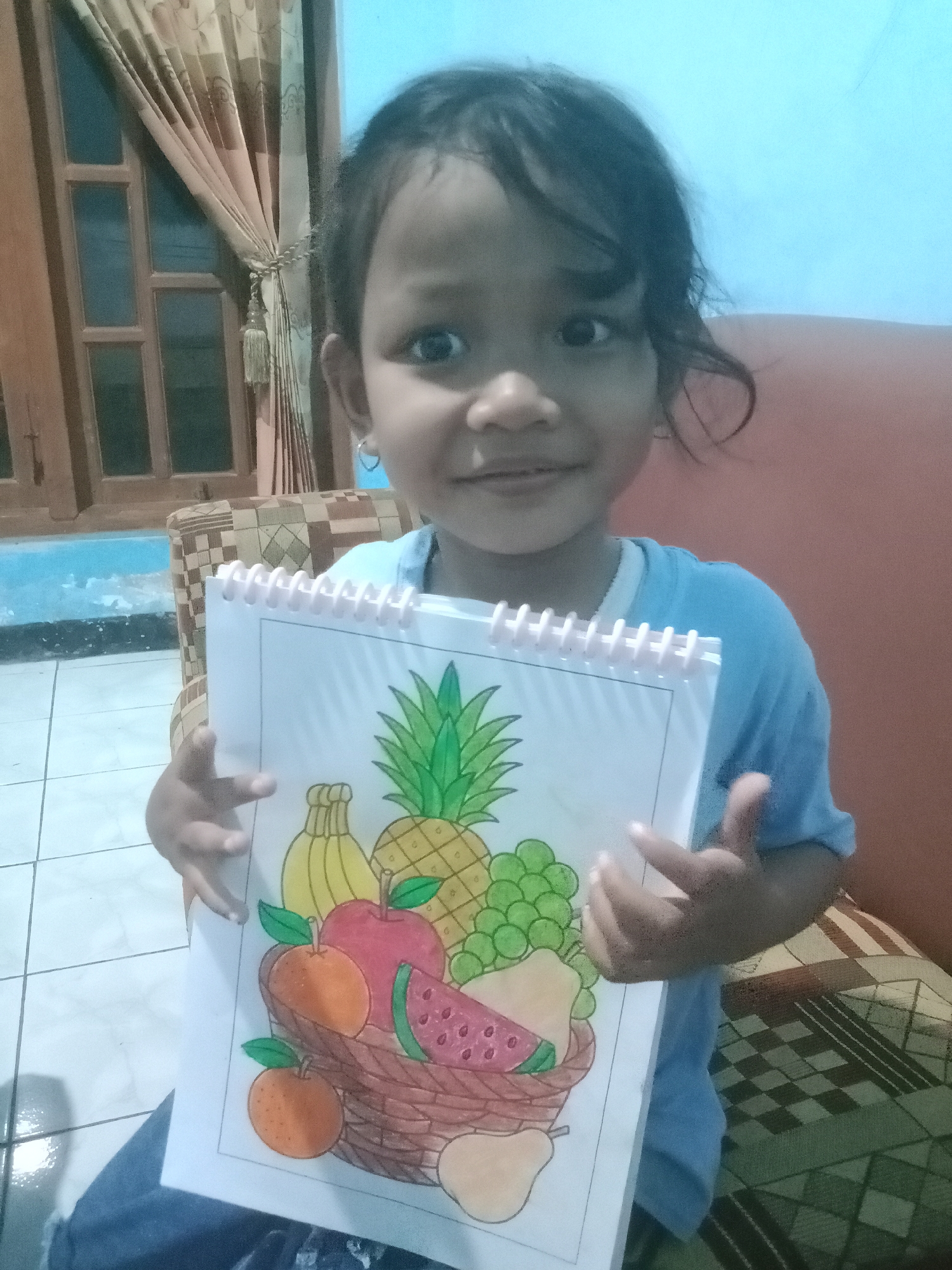 A young girl with dark hair and dark eyes is holding up a drawing of a fruit basket. She is wearing a light blue t-shirt and has a small hoop earring in her left ear. The drawing is in a spiral-bound notebook and depicts a variety of fruits, including a pineapple, bananas, grapes, an apple, oranges, watermelon, and pears, all contained within a woven basket. The background shows a tiled floor, a wooden door with curtains, and a patterned couch. The lighting suggests an indoor setting.
