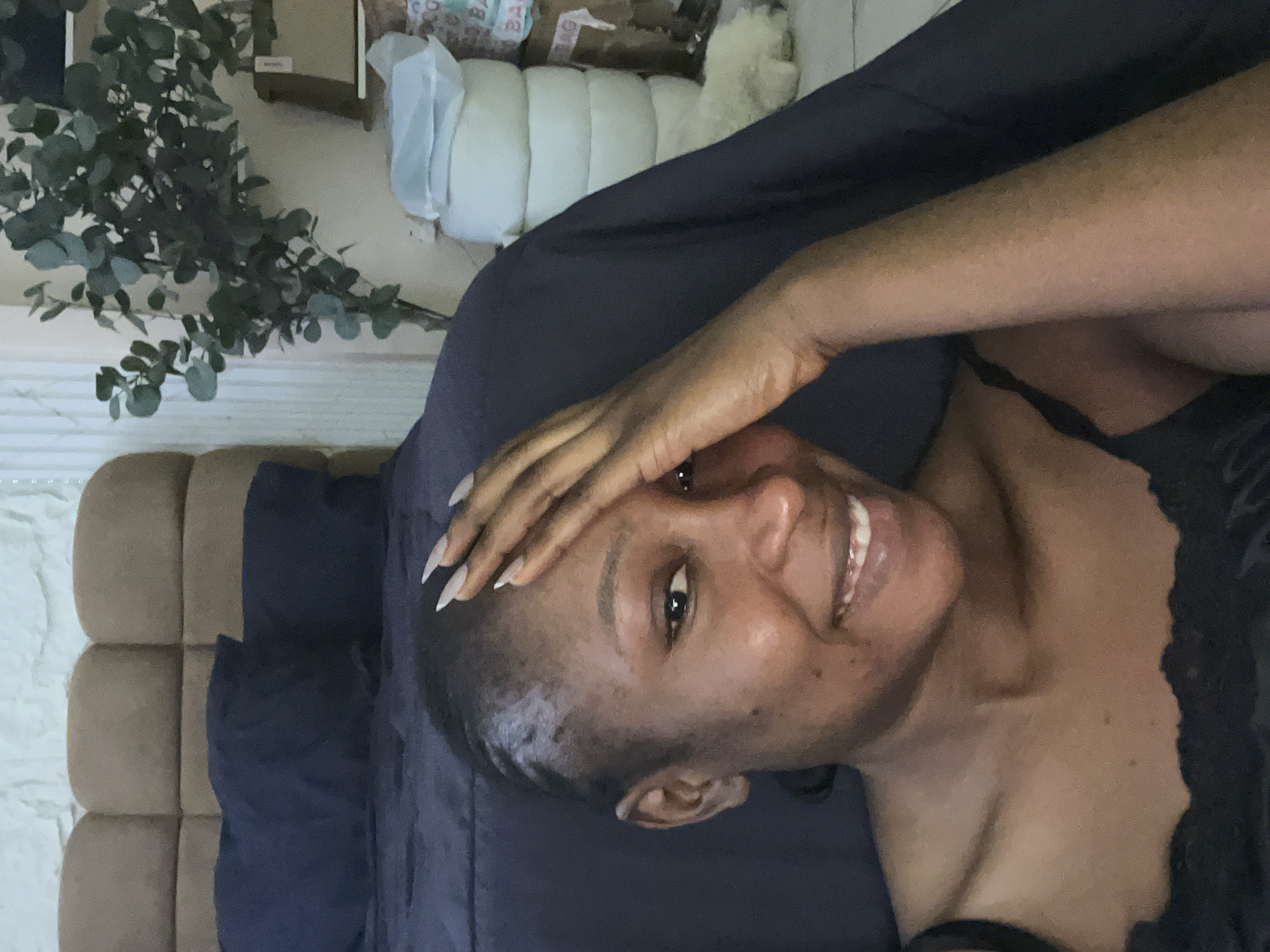 A woman of African descent with dark skin, long fingernails, and dark hair pulled back is shown from the chest up, resting her forehead with her right hand. She is smiling and looking towards the camera. She is wearing a black camisole. The background shows a bed with dark blue pillows and a brown upholstered headboard, as well as a plant with green leaves and stacked white cylindrical objects, possibly mattresses or cushions, and cardboard boxes. The scene is indoors, likely a bedroom. The lighting appears to be natural or soft artificial light. This image is not set in Abuja, Nigeria; it is a generic indoor scene.