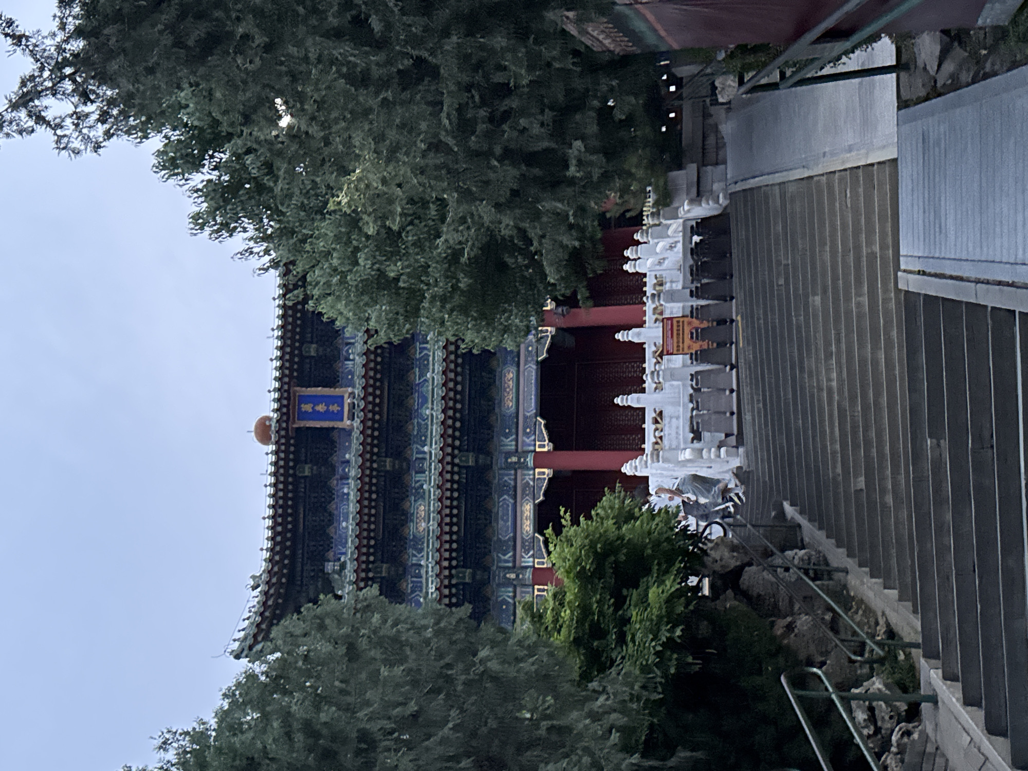 The image depicts a traditional Chinese building partially obscured by trees, viewed from a low angle looking up. The building has a multi-tiered roof with blue and black decorative elements, red columns, and a blue sign with gold lettering in Chinese characters. A person is visible on the stairs near the building, which has white balustrades. The stairs are concrete and lead upwards, alongside a metal railing. The environment appears to be a park or garden setting in daylight. Based on the style of architecture, the building is likely located in the Xicheng District, China.