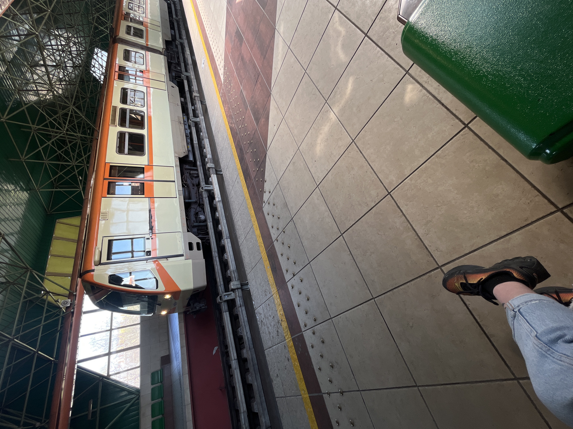 The image depicts a train at a station. A beige and orange train is positioned alongside a platform. The platform has a yellow safety line and a red section closest to the train. The tiles on the platform are mostly beige. A green bench is visible on the platform. A person wearing jeans and black and orange sneakers is sitting on the bench. The image appears to have been taken at an angle and has been rotated counterclockwise. The location may be a subway or metro station in Sofia, Bulgaria.