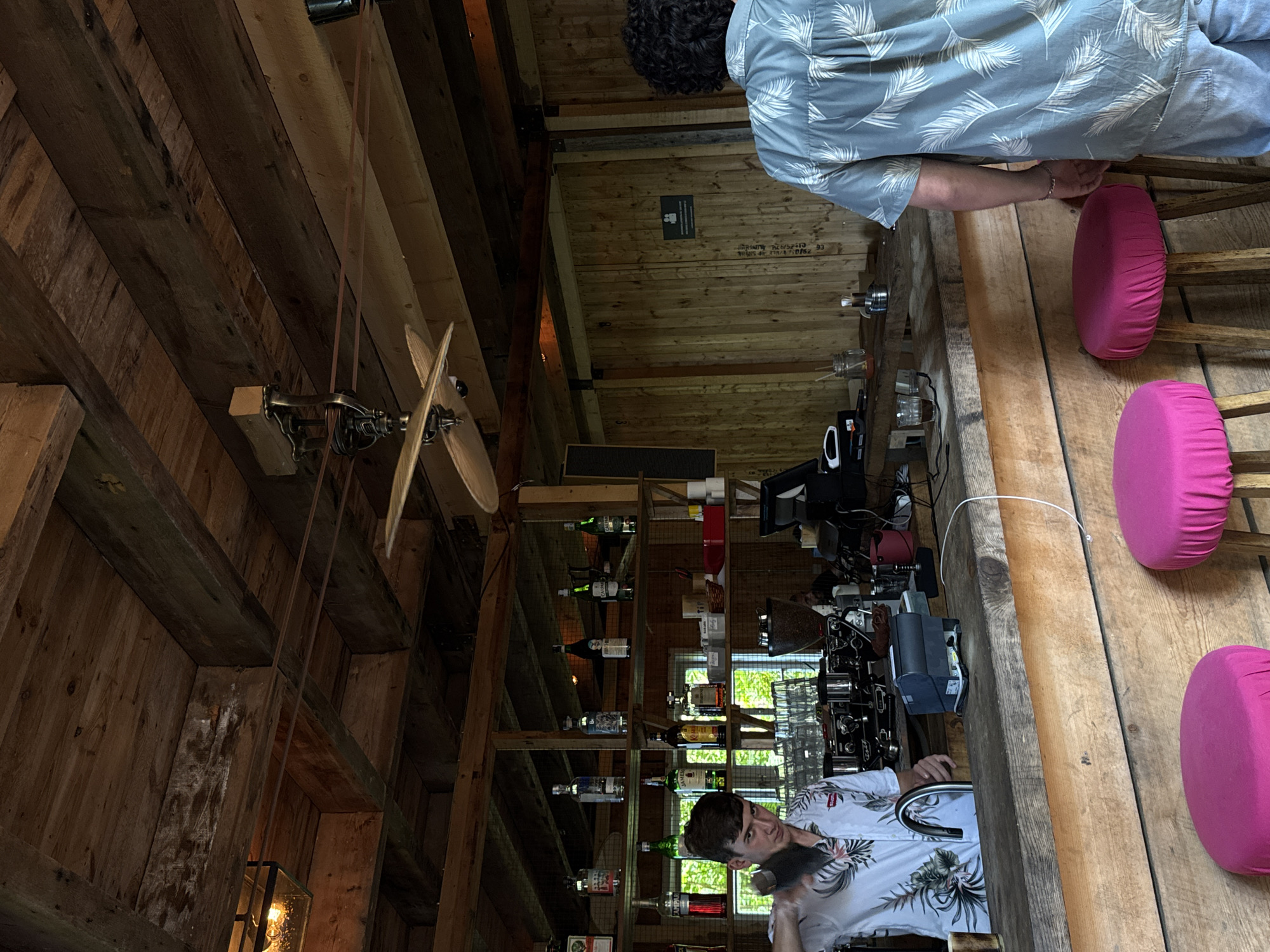 A male bartender with short brown hair is preparing drinks behind a wooden bar. He is wearing a white short-sleeved shirt with a floral pattern. The bar is stocked with various bottles of liquor, glasses, and a coffee machine. In the background, a man with curly dark hair stands facing away from the camera, wearing a light blue short-sleeved shirt with a palm leaf pattern. The setting appears to be an indoor bar with a rustic wooden interior, including exposed beams on the ceiling and wooden walls. The bar is lined with three pink bar stools. The scene is indoors, possibly in the city of Buknari, Georgia, though this is not explicitly stated in the image.