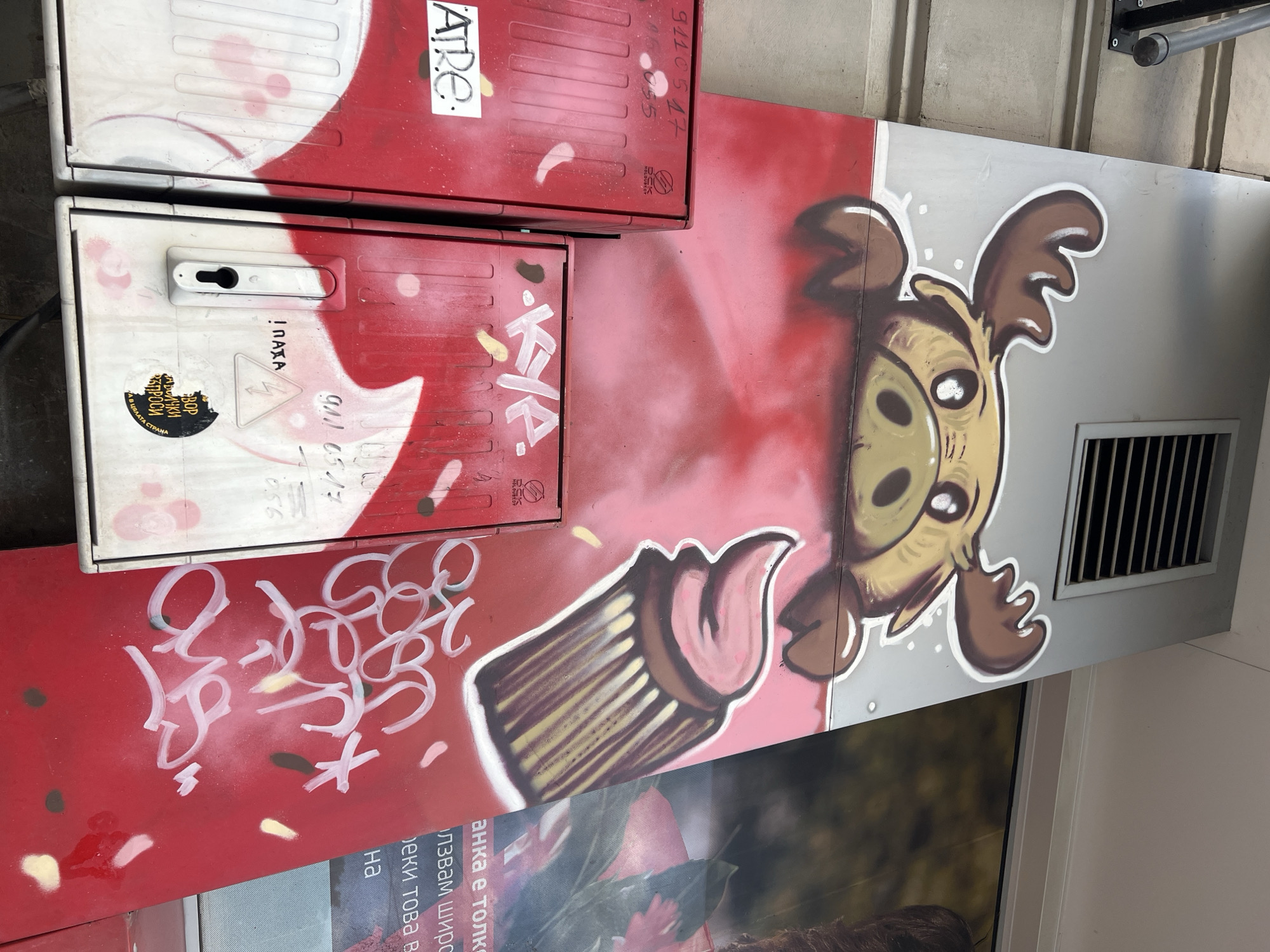 This outdoor shot features a variety of urban elements. On the left, there are two red and white utility boxes with graffiti on them. The upper box has a handwritten "ATRE" and some numerical markings. The lower box has a handle, a yellow sticker, and more graffiti, including the words "! ΠΑΤΑ" and some numbers. A sticker on the lower box reads "BOP НА ВСИЧКИ ВЪПРОСИ В ЦЯЛАТА СТРАНА". To the right, there's a large mural that appears to be on the side of a building. The mural is a mix of colors, with a cartoonish moose holding a cupcake in the design. Parts of the mural are obstructed by what appears to be a poster or advertisement for the brand 'Lidl'. The location is Sofia, Bulgaria.