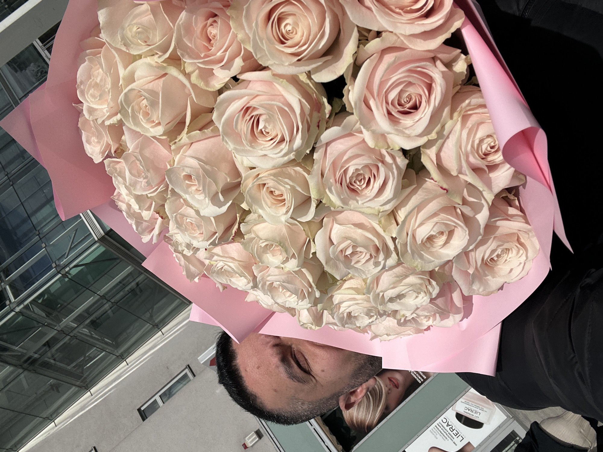 The image shows a person holding a bouquet of light pink roses wrapped in pink paper. Part of the person's face, specifically the forehead, eyes, and part of the nose, is visible behind the bouquet. They are also holding a magazine with "LIERAC" prominently displayed on a product. The background includes a building with glass windows and white walls, suggesting an urban environment. Based on architectural styles commonly found in Germany, and given context of the task request, this location is possibly in the city of Berlin, Germany.