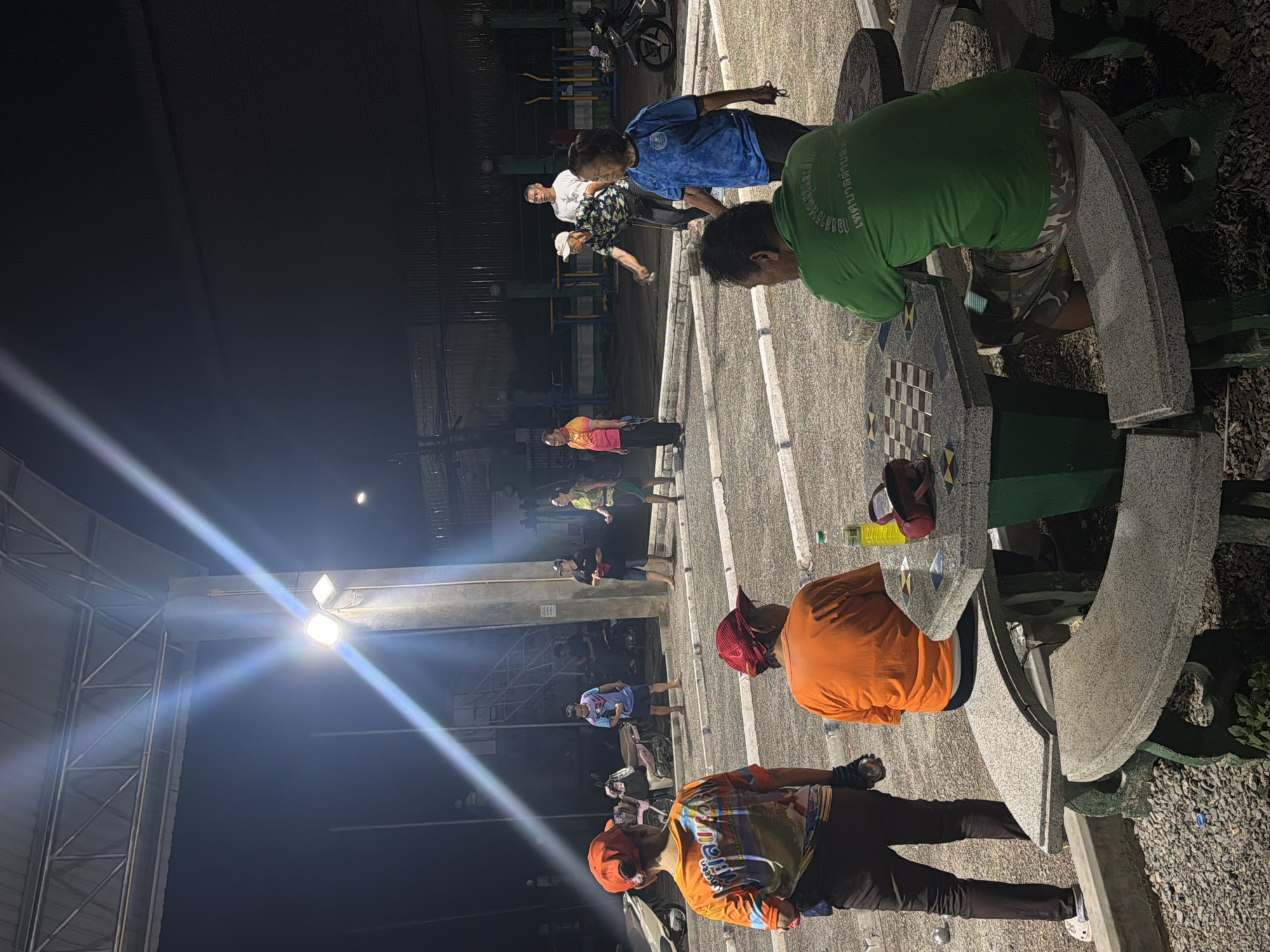 At night, in Chang Phueak, Thailand, a group of people are engaged in various activities. Several individuals stand near a walkway under bright lights, while others are gathered around tables with chessboards. One man is seated at a table, wearing an orange shirt and red cap. Another man in an orange patterned shirt and brown pants stands nearby. A few more people are further down the walkway. A motorcycle is parked on the walkway. The environment consists of an open, illuminated area with a paved walkway.