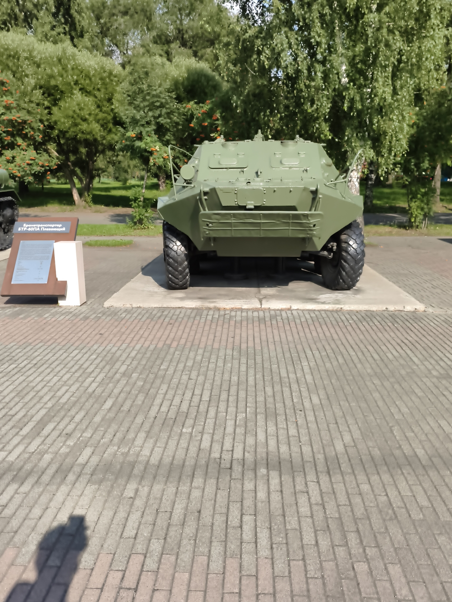 A green armored personnel carrier is displayed outdoors on a paved area. The vehicle is the primary subject, positioned in the center of the frame and viewed from the rear. To the left of the APC, a display board with text and a white pedestal are visible. The background consists of lush green trees and foliage, suggesting a park or outdoor exhibition space. The ground is paved with interlocking bricks, and a shadow of a person holding a camera is cast on the ground in the lower left portion of the image. The scene appears to be a sunny day. The APC is likely part of a museum exhibit, given the accompanying display. This location is the "Place of Memory" memorial complex in Victory Park, Chelyabinsk, Russia, specifically honoring military vehicles.