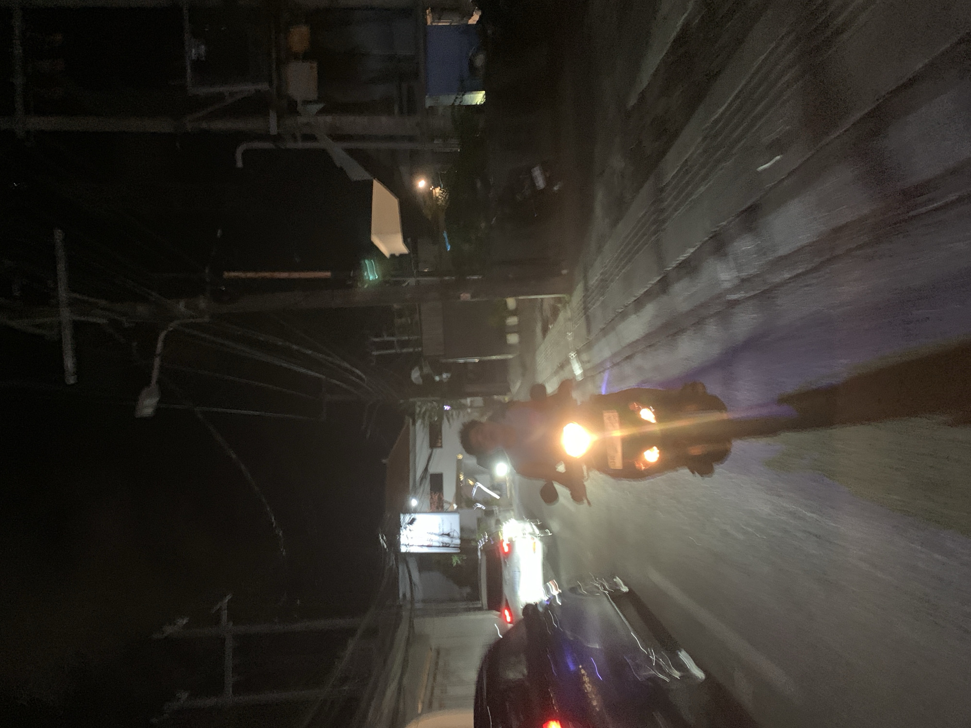 At night in North Kuta, Indonesia, a person rides a motorcycle with its headlight illuminating the wet asphalt road. There is a car behind the motorcycle. The area is urban with buildings, telephone poles, and wires visible along the road.
