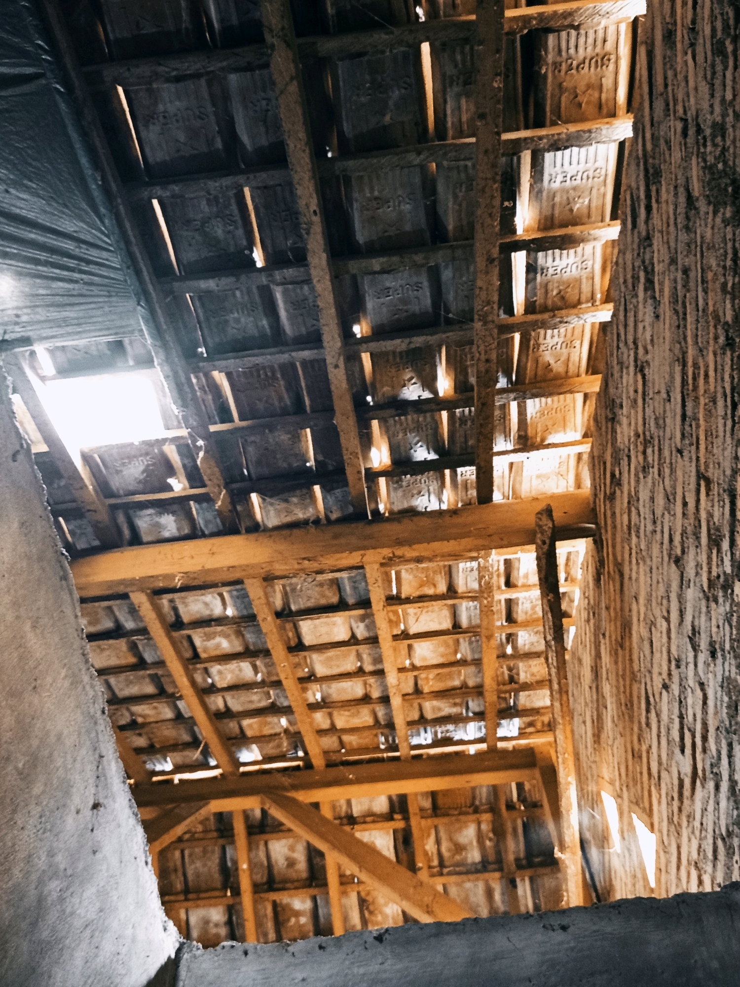 The image depicts the underside of a damaged roof structure in what appears to be an unfinished or derelict building. The roof is composed of wooden beams and supports, with rows of what seem to be clay tiles visible from below. Many of the tiles are marked with the word "SUPER" in raised lettering, suggesting they are commercial roofing tiles. Gaps between the tiles allow shafts of light to penetrate, indicating holes or missing sections in the roofing material. A large section of grey, rough-textured material, possibly concrete or plaster, occupies the left side of the foreground, obscuring part of the roof structure. The overall impression is one of disrepair and neglect. The location is identified as Karang Sari, Indonesia. There are no people or active interactions visible in the image.