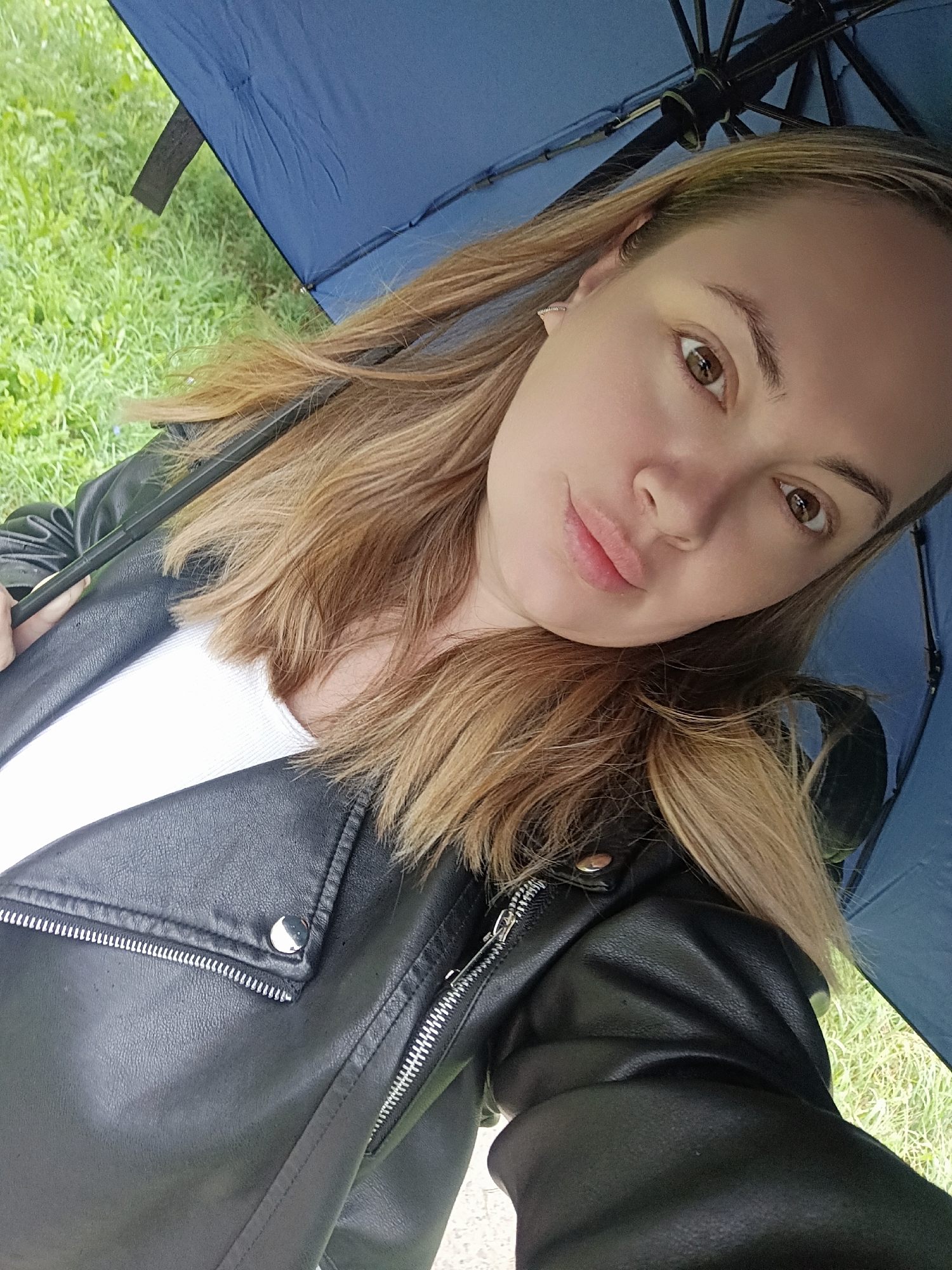 A young woman with blonde hair and brown eyes is captured in a selfie under a blue umbrella. She is wearing a black leather jacket over a white top. The background consists of green grass and what appears to be foliage. The scene is outdoors, likely in a park or garden, and the overcast sky suggests it might be raining or has recently rained.