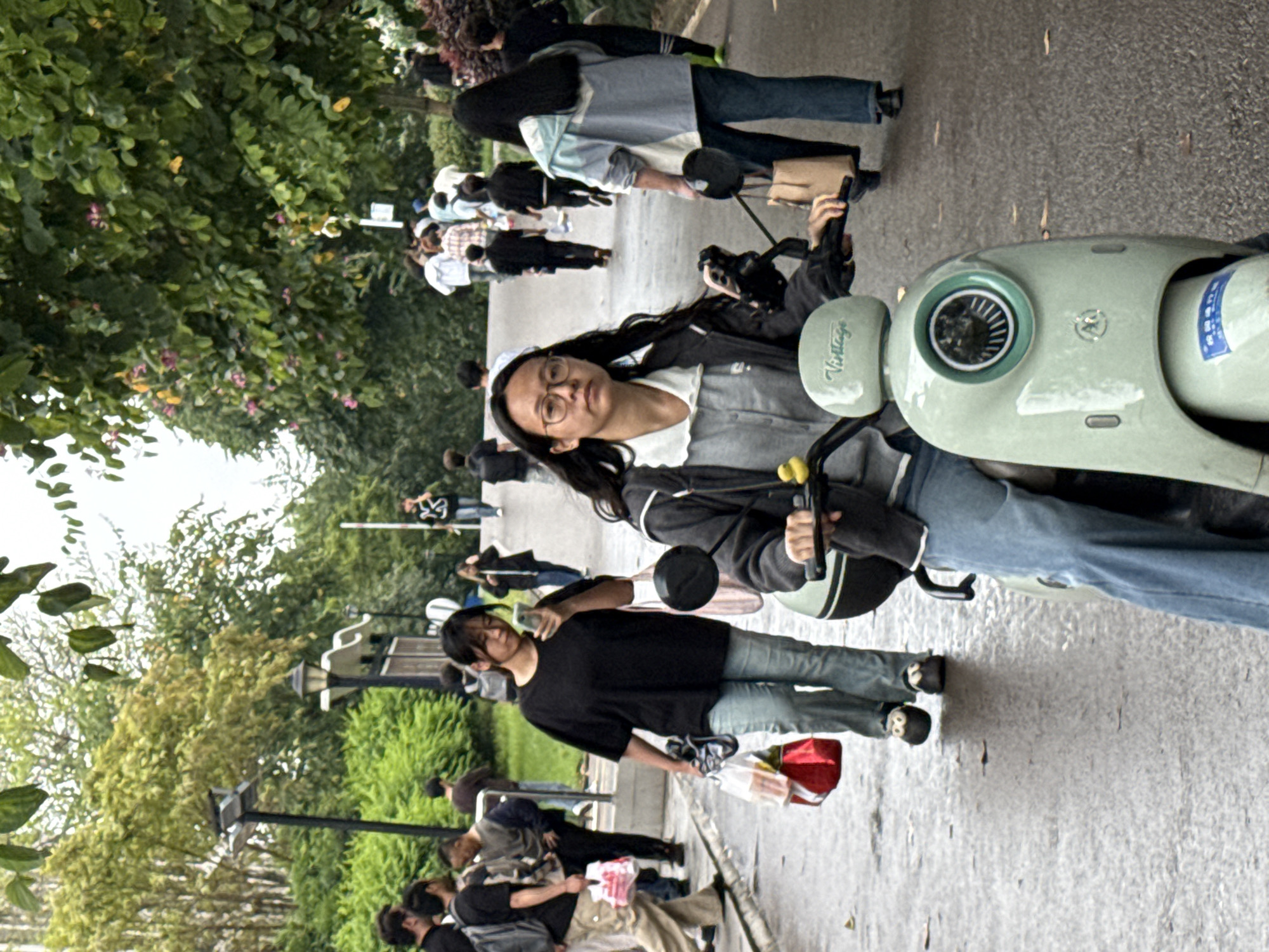 The image depicts a street scene in Jiangjin, China. An Asian woman wearing glasses and a grey jacket over a white shirt is riding a light green electric scooter on a paved road. Behind her, another person is walking on the side of the road. Several other people are visible in the background, along with trees and street lights.