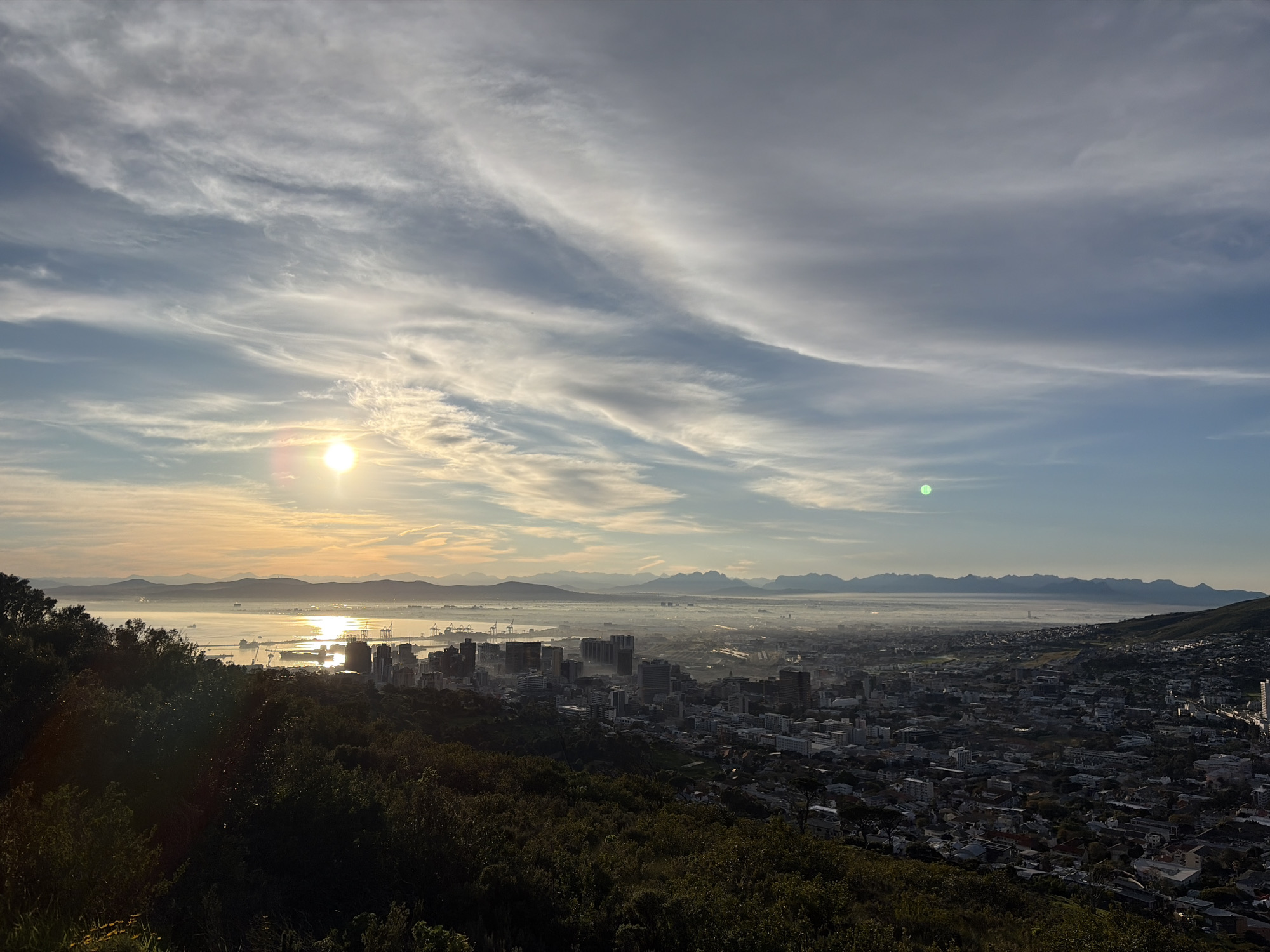 The image shows a panoramic view of Cape Town, South Africa, from a high vantage point, likely Signal Hill or Lion's Head, overlooking the city and Table Bay. The sun is low in the sky, casting a warm glow and long shadows, suggesting it is either sunrise or sunset. Wispy clouds streak across the sky, illuminated by the sun.
In the foreground, a dense cluster of dark green trees and foliage covers the hillside. Beyond the trees, the city of Cape Town unfolds. Numerous buildings, including high-rises and residential areas, stretch out towards the coast. The urban landscape transitions into the harbor area, where cranes and industrial structures are visible. Table Bay is a calm expanse of water, reflecting the sunlight and dotted with ships and boats. In the distance, a range of mountains forms the backdrop to the city, shrouded in a light haze. The overall impression is a serene and expansive cityscape bathed in the soft light of either dawn or dusk.