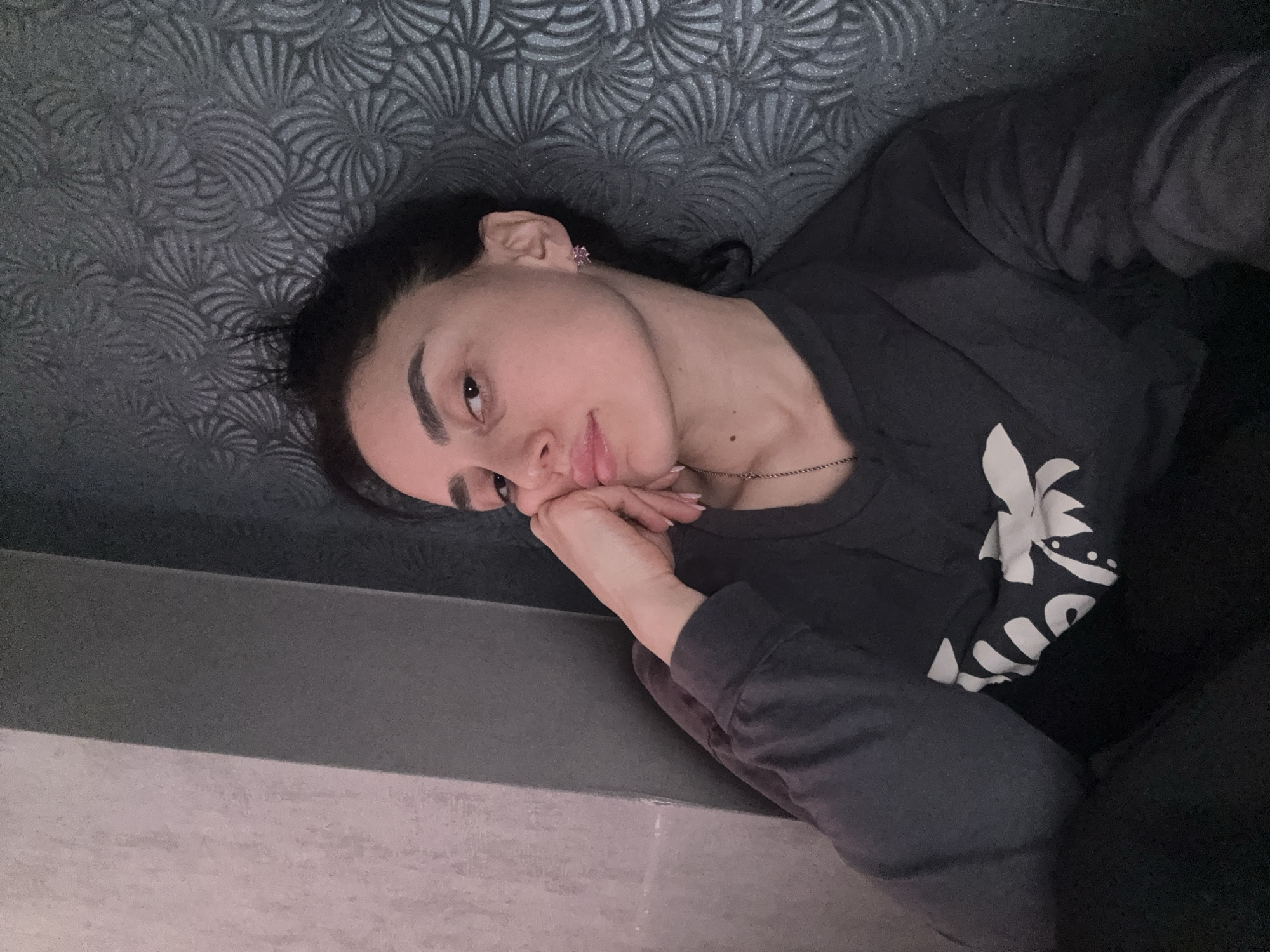 A woman lies on a patterned grey upholstered headboard or wall. She is wearing a dark-colored long-sleeved shirt with a white floral design on the sleeve. She rests her head on her hand, looking at the camera. She wears a necklace and earrings. The lower part of the frame shows the side of a gray or light-colored surface. The location is unidentifiable from the image, and Tbilisi, Georgia cannot be confirmed.