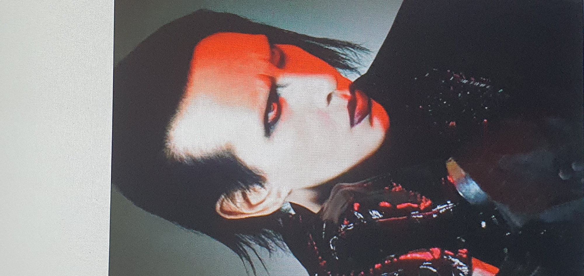 The image is a close-up portrait of a person with pale skin, black hair, and dark eye makeup. They are wearing a black, high-collared garment, possibly a jacket or coat, with a textured, possibly reflective, surface on the left side. Red lighting illuminates the right side of their face, creating a strong contrast. The person is looking downwards and slightly to their right. The background is out of focus and appears to be a solid, neutral color. There is no indication of the city of Bat Yam, Israel, or any other specific location within the image.