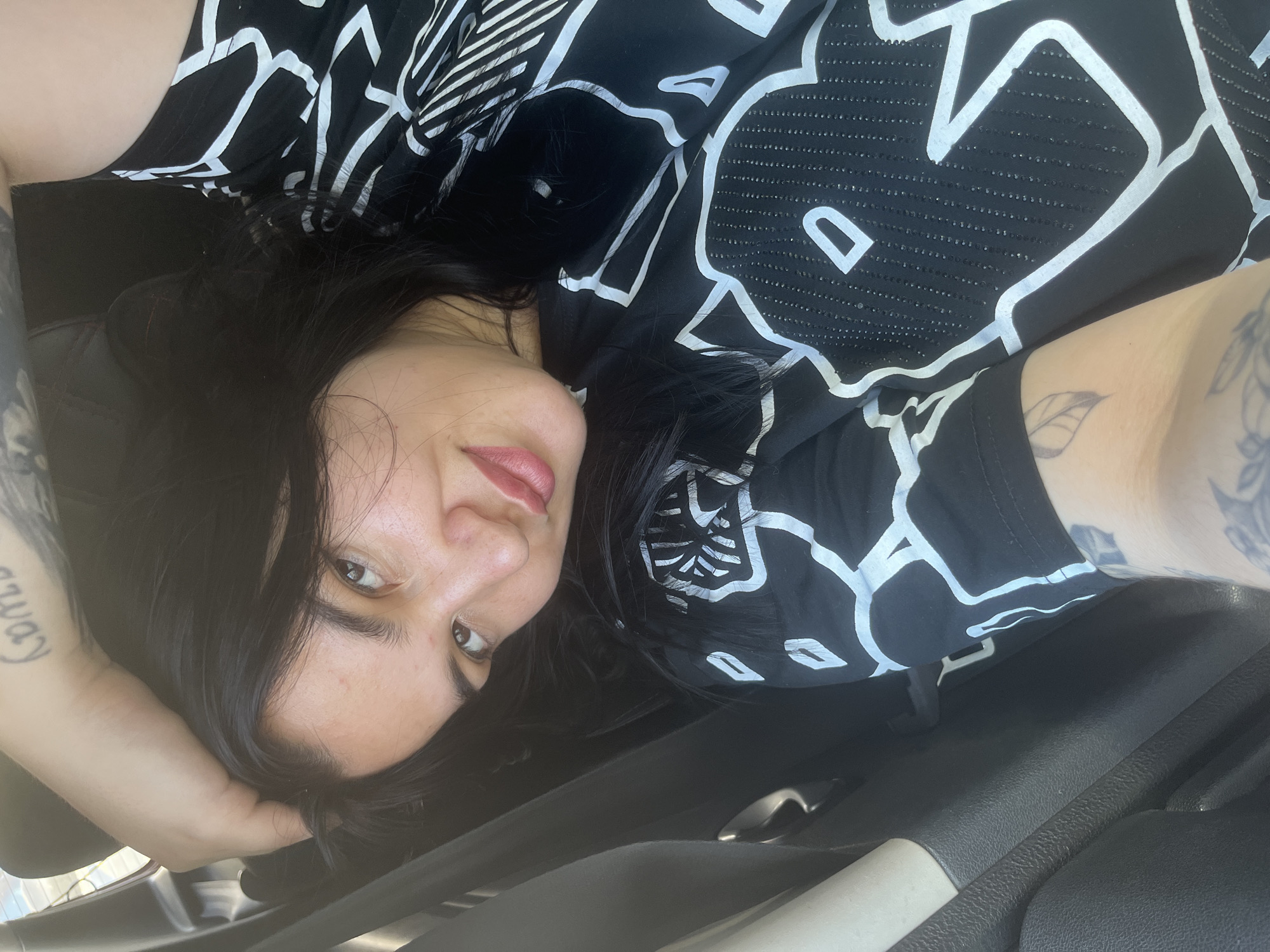 A woman with dark hair and light skin is lying down, looking at the camera. She is wearing a black and white patterned shirt. Her left hand is behind her head, and her right arm, which is tattooed, is visible. The interior of what appears to be a car is visible in the background, including a car door handle. The location is potentially a car in Chaikovsky, Russia.