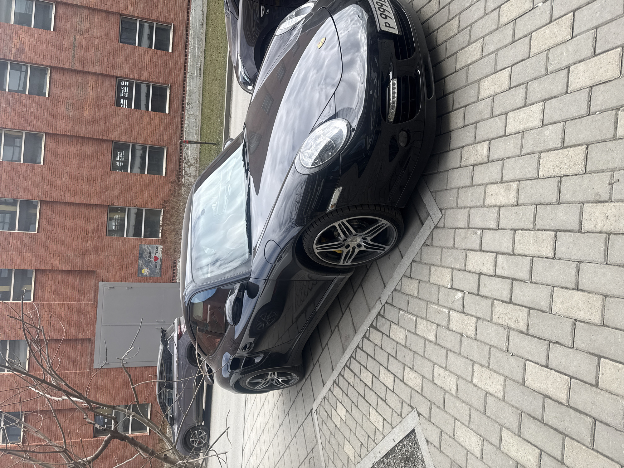 The image shows two parked cars on a brick paved area. The foremost car is a black Porsche with a license plate beginning with "P 999". The car next to it is dark grey. Behind the cars is a red brick building with windows. In the lower left corner of the image, there is a bare tree branch. Based on the license plate of the Porsche, this image was taken in Yekaterinburg, Russia.