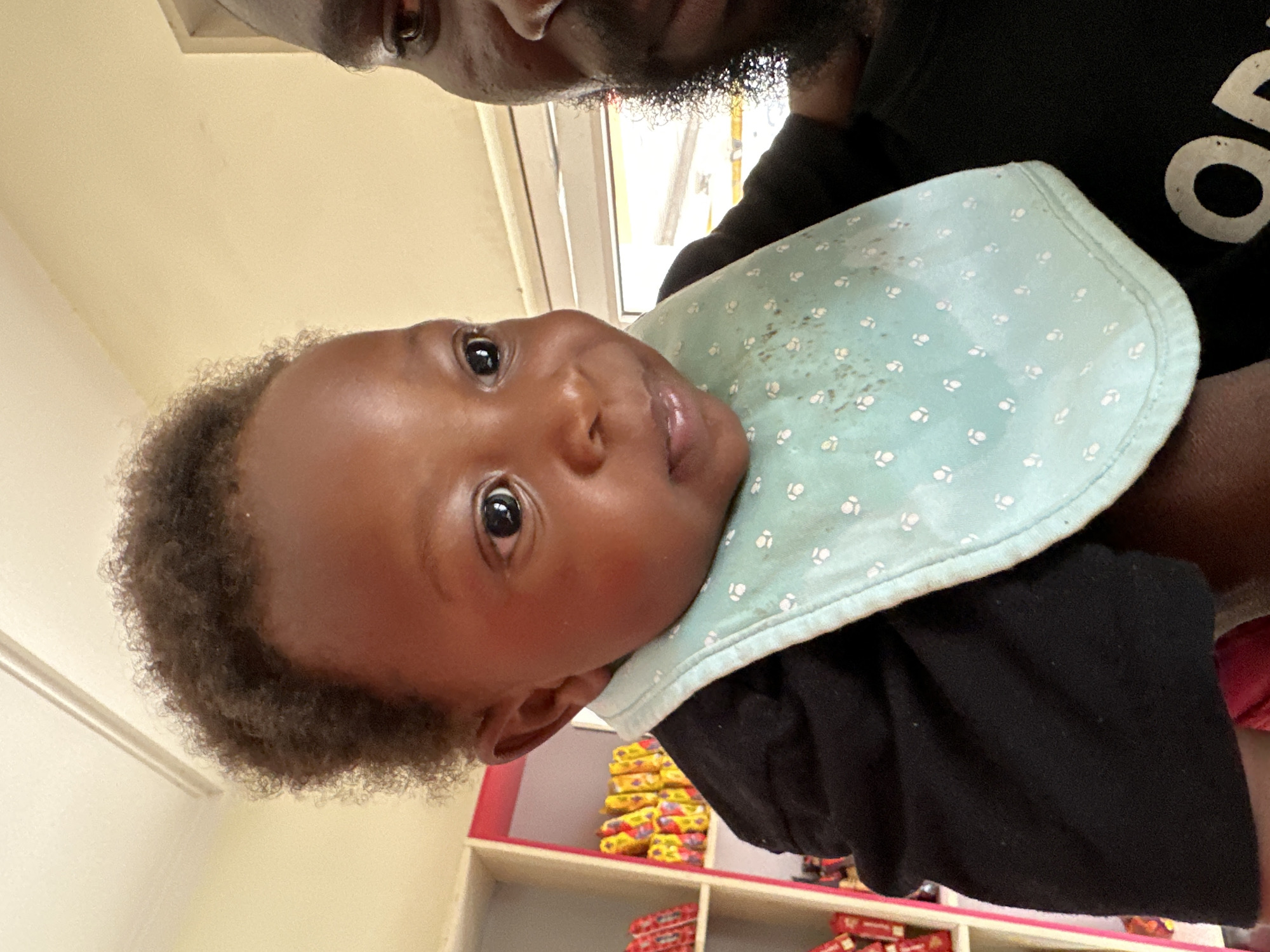 A close-up, vertically oriented image, originally rotated 90 degrees counter-clockwise, captures an adult holding a baby indoors.
The baby, centrally positioned and looking directly at the camera, has dark skin, dark curly hair, and large, expressive dark eyes. Their expression is one of curiosity or a slight smile. They are wearing a light blue bib with a pattern of small white dots, which shows some faint dark stains.
Partially visible in the upper portion of the frame, behind the baby, is an adult. Only the adult's lower face, featuring a beard or stubble, and the upper part of their body are seen. They are wearing a black t-shirt, on which a partial white letter, resembling an 'o', is visible.
The background reveals an indoor setting. A bright window is visible towards the upper left, indicating daytime. Below the subjects, shelves are stacked with various packaged items, including bright yellow and red packaging, suggesting a retail environment such as a small shop or kiosk. The scene conveys a tender, everyday moment between the adult and the baby.