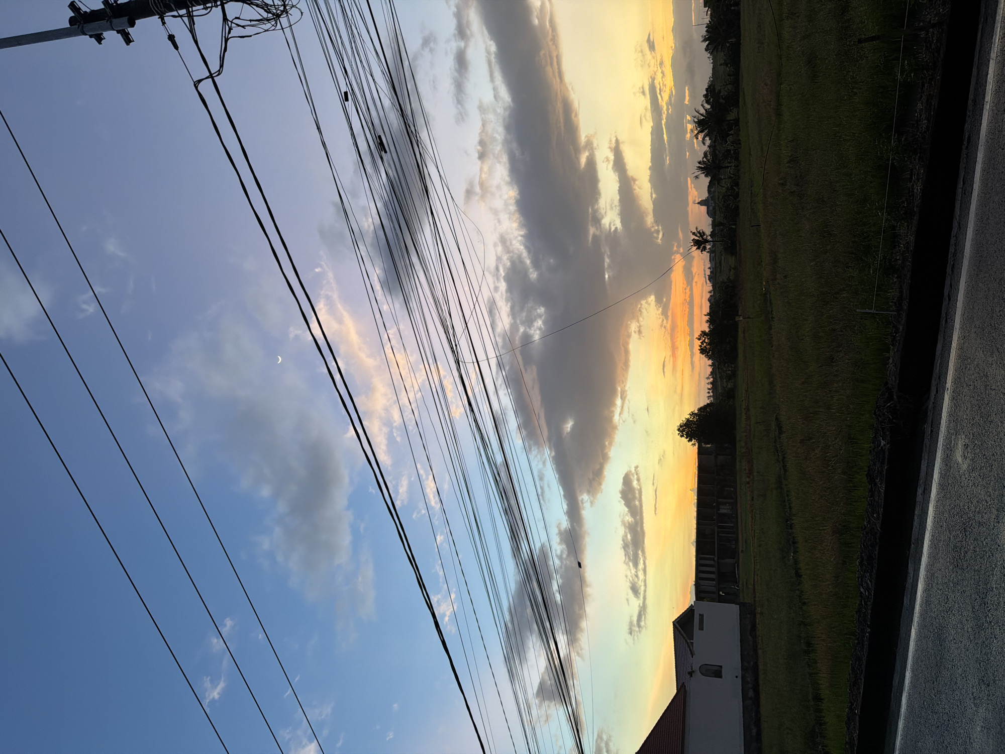 The image captures a sky at twilight in Mengwi, Indonesia. The sky transitions from blue with scattered clouds to orange and yellow hues near the horizon. A crescent moon is visible in the blue portion of the sky. Overhead power lines crisscross the view. The foreground shows a rice paddy, a building with a red tiled roof, and a section of road.