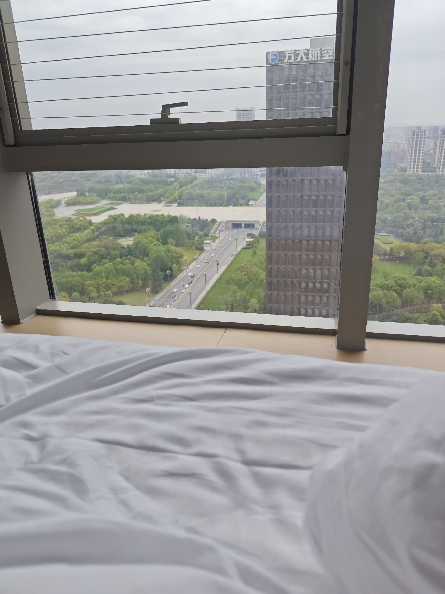 The image depicts an interior view from a high-rise building. In the foreground, a rumpled white bed sheet and a partially visible white pillow are in focus, suggesting a scene from a bedroom or hotel room. The main subject is the view from a window, which looks out upon a cityscape.
Through the open window, a river and a wide roadway are visible, surrounded by green trees and parkland. Several cars are moving on the road. A tall, modern building is situated close to the window, partially obstructing the view. The upper portion of another skyscraper is visible, bearing a logo with Chinese characters and what appears to be the English text "Great Aircraft."
The scene is set in Liangjiang, China, as indicated by the name on the skyscraper. The weather appears overcast or cloudy, casting a diffused light on the outside scenery. The image presents a still, indoor scene juxtaposed against an urban outdoor landscape.