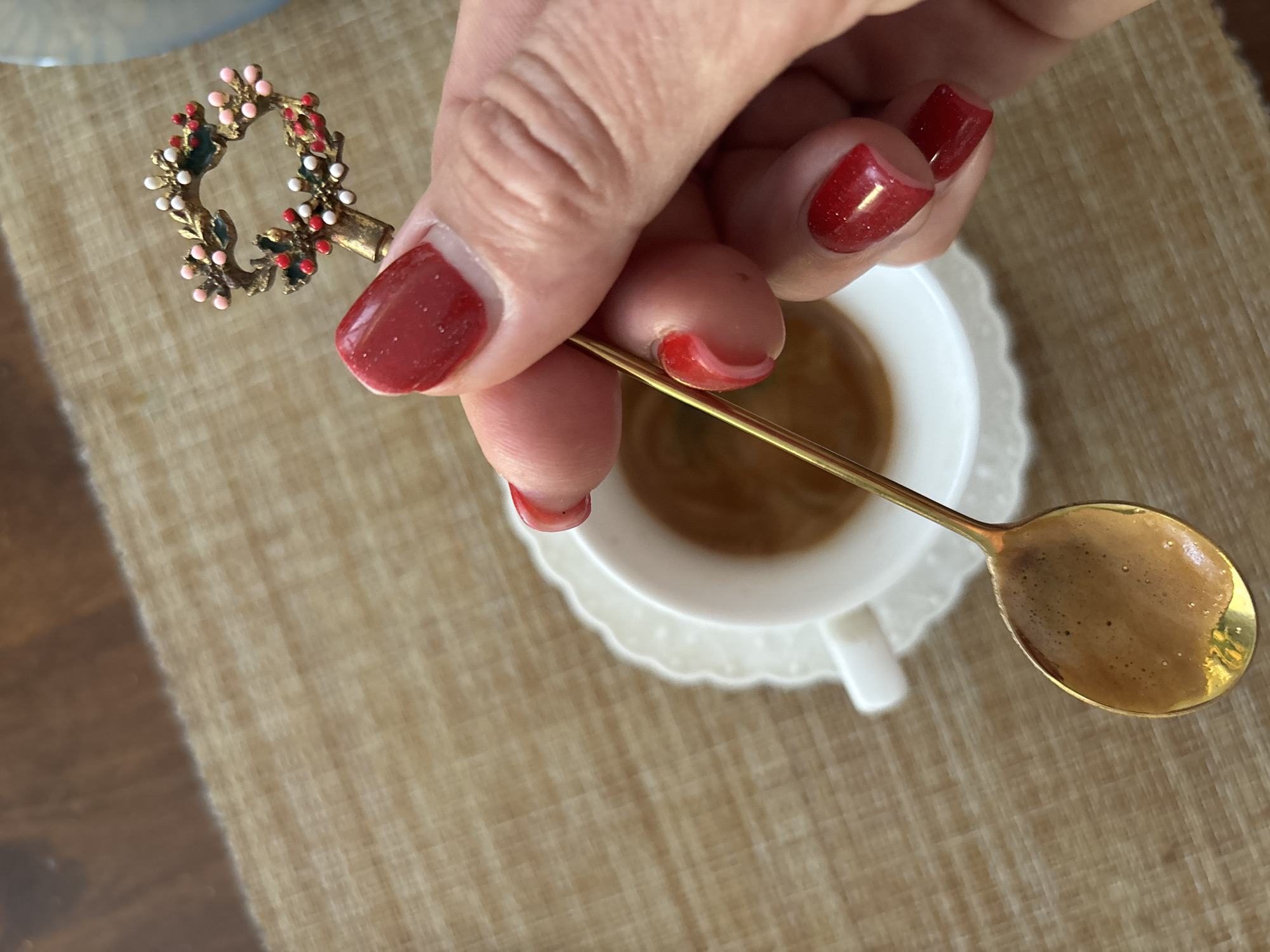 An overhead, slightly angled shot shows a person's right hand holding a gold spoon with a decorative festive wreath on its end. The spoon is positioned above a white teacup filled with coffee. The background consists of a textured brown placemat and a wooden surface. The person's fingernails are painted a bright red. The image does not contain any information about the city of Sochi, Russia.