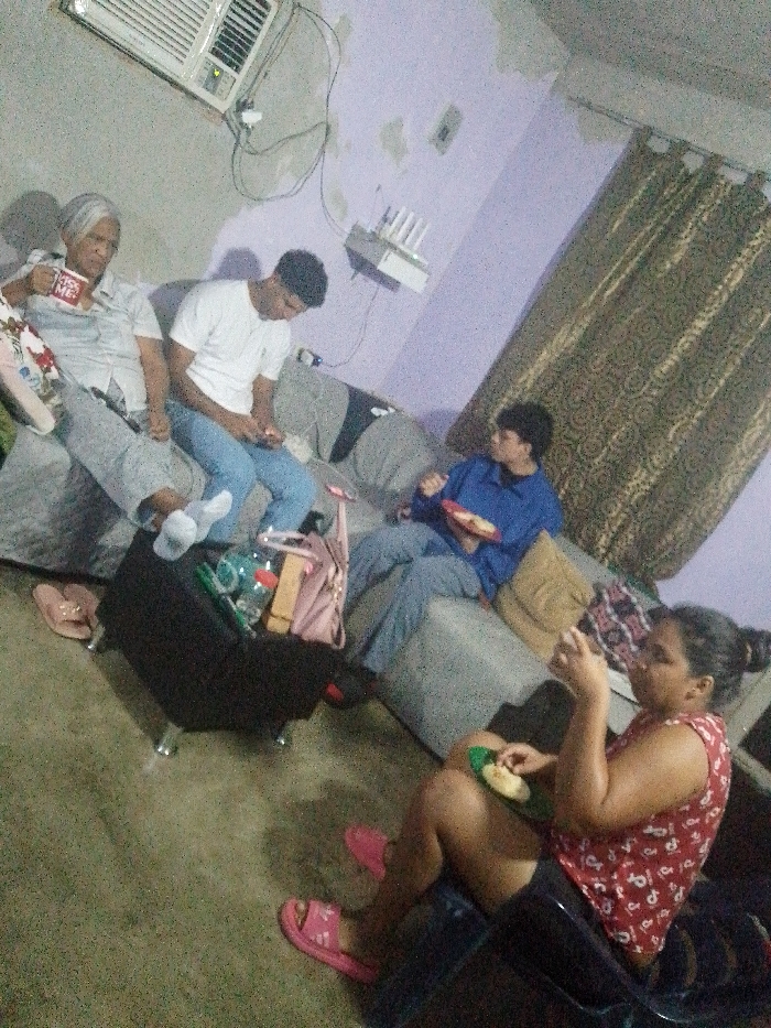 Four people are relaxing indoors. An elderly woman holds a mug on the far left. A young man in a white shirt looks at his phone. Another person in a blue shirt eats from a plate. In the foreground, a woman in a red patterned top also holds food. They sit on a gray sofa near a black suitcase with items inside. The room has brown curtains and light purple walls. There's an air conditioner and a router mounted high on the wall.