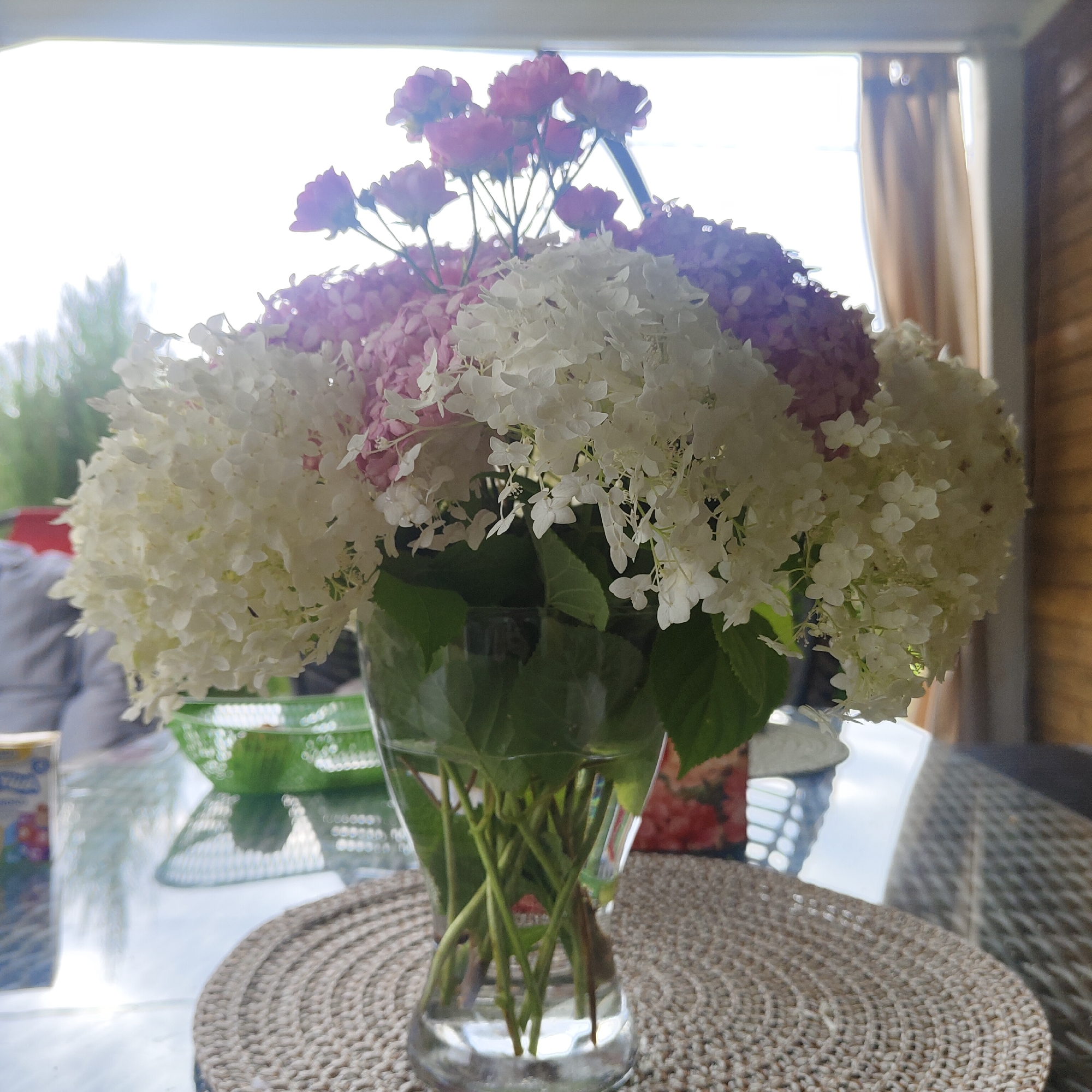 A bouquet of pink and white hydrangeas and roses is displayed in a clear glass vase on a textured, woven placemat atop a glass table. The bouquet is large and fills a significant portion of the frame. Behind the vase, a bright window with sheer curtains is visible, suggesting an indoor setting, possibly on a patio or sunroom, with sunlight streaming through. In the background, elements of outdoor greenery and furniture can be seen, along with a woven chair. To the left of the vase, a portion of a box with colorful graphics is visible. The overall scene depicts a decorative floral arrangement indoors, with natural light playing a significant role. The location "Берега Днп, Russia" is not visually discernible from the image itself.