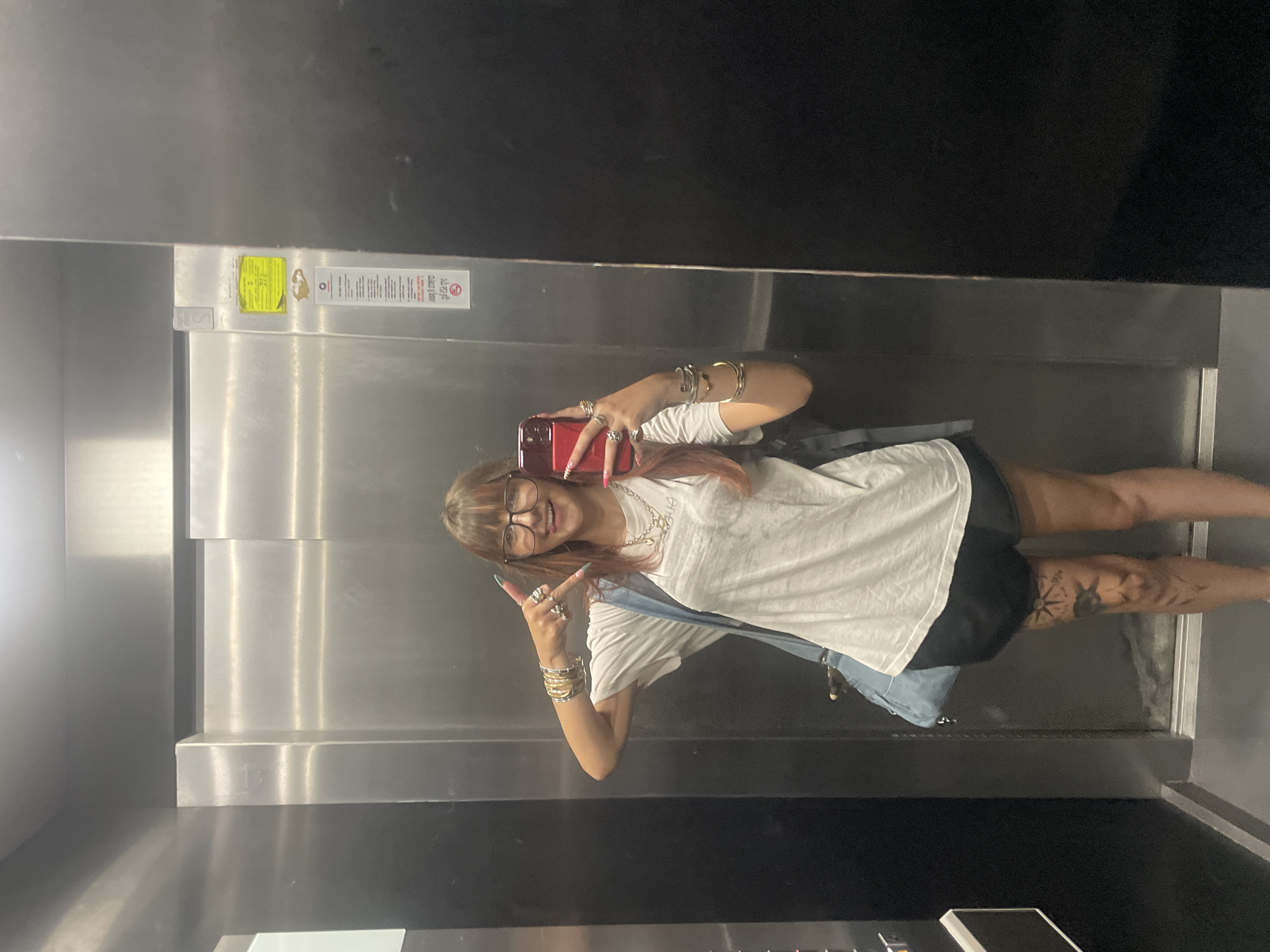 A young woman with reddish-brown hair and bangs, wearing glasses and a white t-shirt, is taking a selfie in an elevator in Raanana, Israel. She is holding a red smartphone in her right hand and making a peace sign with her left hand. She is wearing several bracelets on both wrists and multiple rings. A denim bag is slung over her left shoulder. She has a tattoo on her right thigh. The elevator has stainless steel walls and doors. A yellow sticker and some text in Hebrew are visible on the elevator wall.