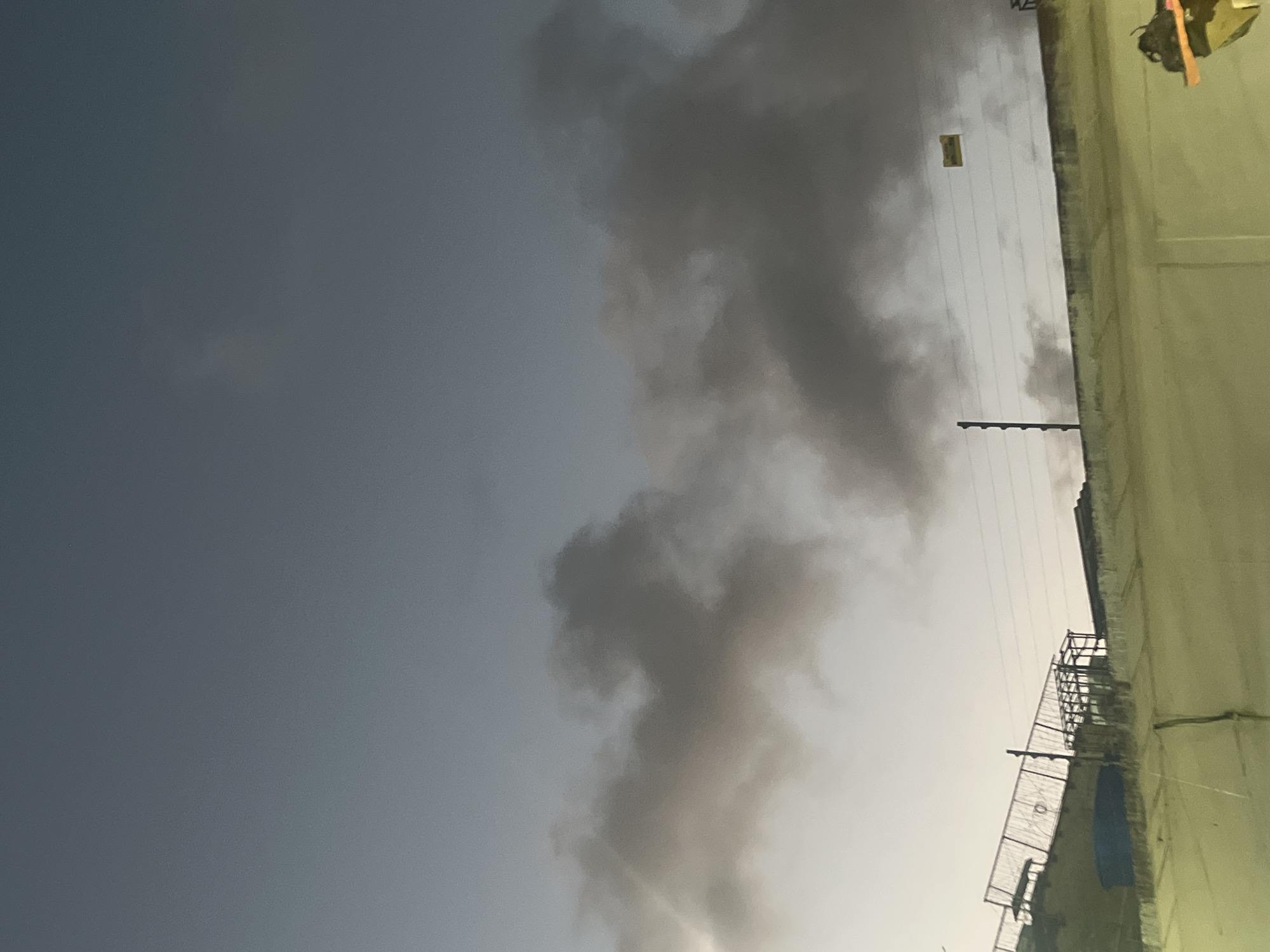 The image shows a partially cloudy sky with dark grey clouds dominating the upper portion. Below the clouds, there is a wall and structures that appear to be part of a construction site or industrial area. A fence with visible wires runs along the top of the wall, and a sign with yellow text is attached to the wires. The lighting suggests it is either early morning or late evening. No people are visible in the image. The overall scene is urban or industrial, with a sense of unfinished construction or an active work area. The location of Conuco Viejo, Venezuela is not discernible from the visual elements in the image itself.