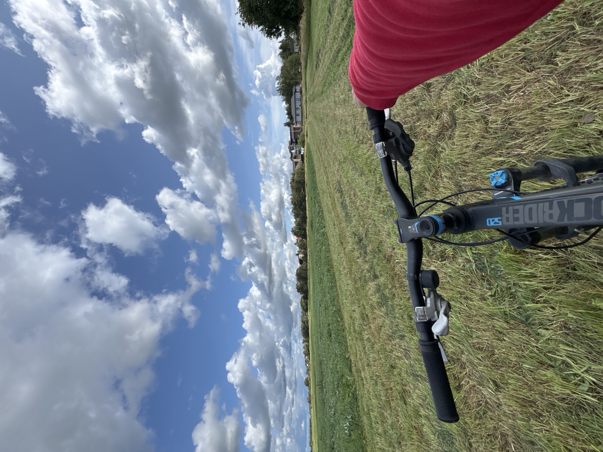 A first-person perspective shows a person riding a black Rockrider 520 bicycle through a grassy field with scattered hay. The rider's right arm, clad in a red sweater, is visible on the handlebar. The background features a line of trees and buildings, possibly houses, under a sky with scattered clouds. The location is identified as Kempelevo, Russia.