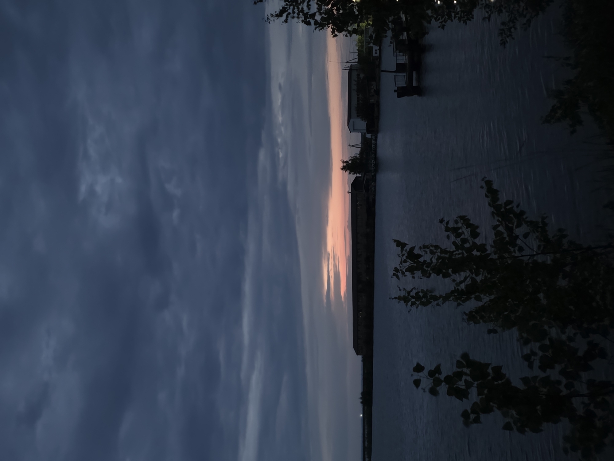 The image captures a twilight scene. A body of water, possibly a river or lake, stretches vertically through the center. The sky is predominantly dark with stormy clouds on the left and a narrow band of pinkish-orange hues along the horizon. There are buildings visible along the shoreline on the right, and vegetation, possibly trees, partially obscures the view from the left and right edges. The scene appears to be in the city of Chaikovsky, Russia.