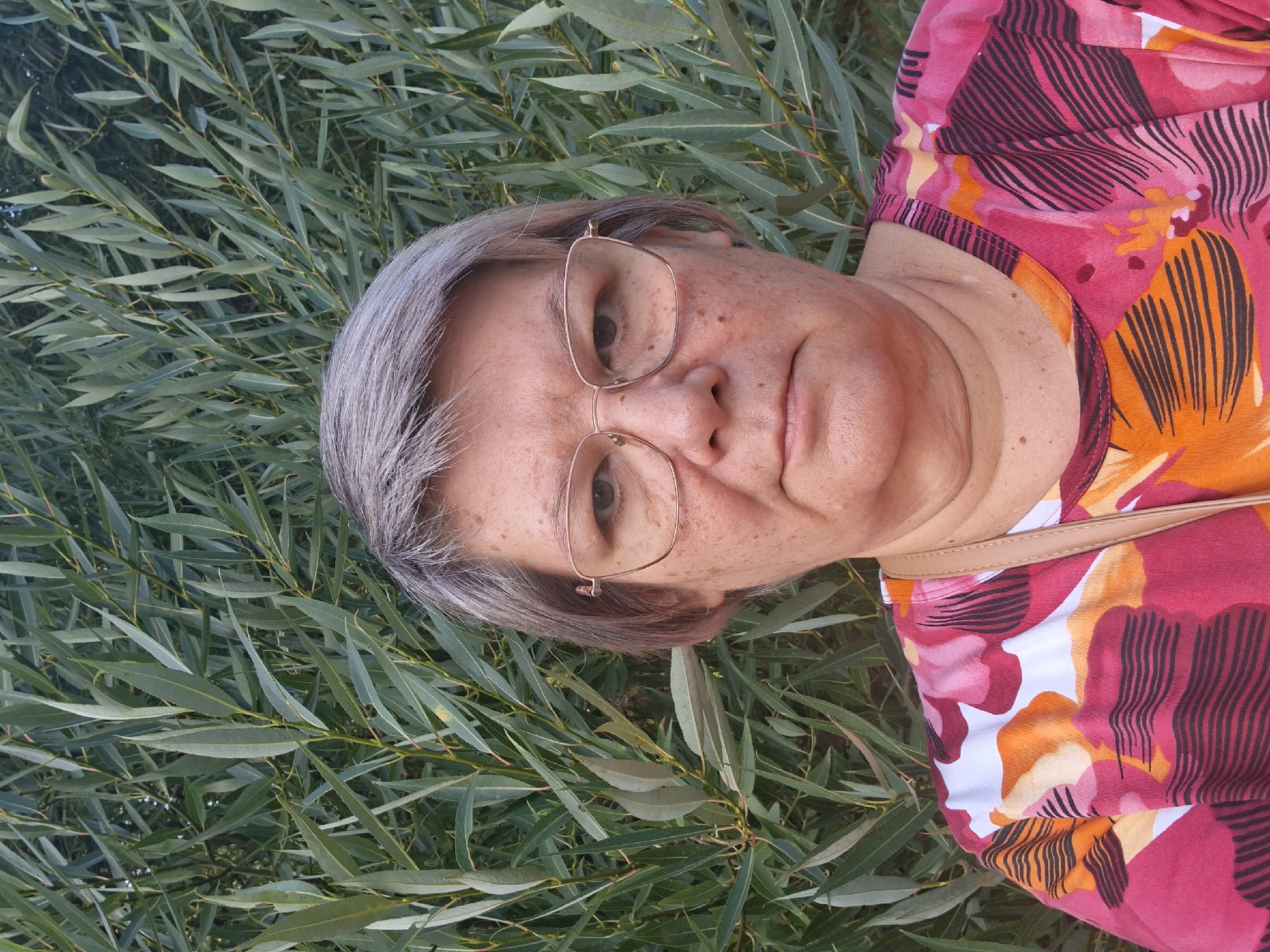 A close-up, head-and-shoulders outdoor portrait of a middle-aged woman with short, grey hair and glasses. She is looking directly at the camera with a neutral expression. Her skin has some freckles and sun spots. She is wearing a colorful, floral-patterned shirt. Behind her is a dense background of green foliage, likely a tree or shrub. The image appears to have been taken during daylight hours. There is no visible indication of the specific location within Novosibirsk, Russia.