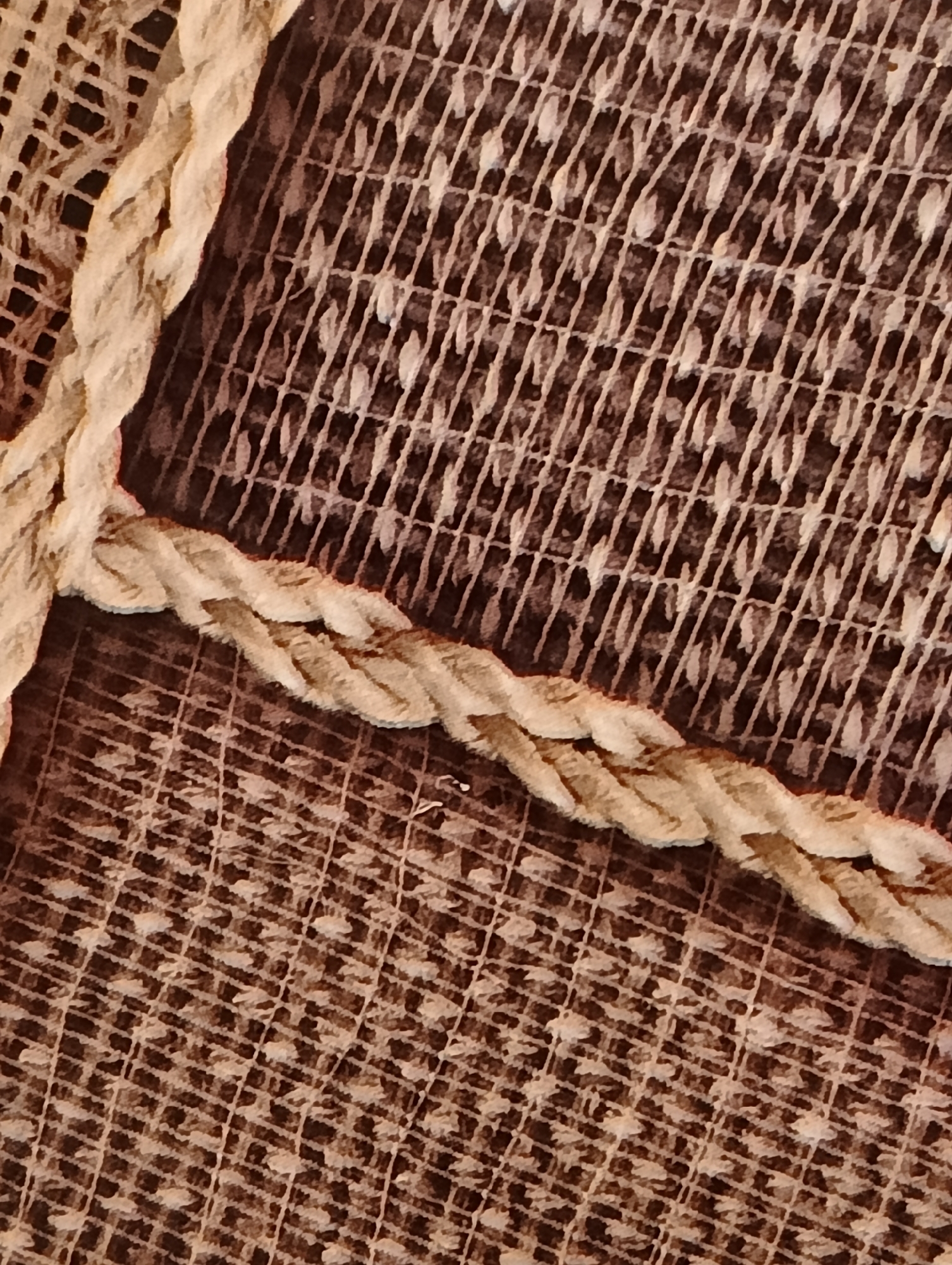 This close-up image, located in the city of Biliaivka, Ukraine, presents a detailed view of two distinct textile elements. A thick, light-beige braided rope, exhibiting a visibly fuzzy texture consistent with natural fibers, runs diagonally across the frame in a gentle 'S' curve. The rope partially overlays a darker, patterned textile background. This background fabric features a grid of fine, light-brown lines, within which numerous small, irregularly shaped, lighter elements resembling leaves or petals are densely arranged, creating a textured, decorative pattern. The scene is devoid of any human presence, subjects, actions, or interactions, focusing exclusively on the tactile qualities and interplay of the textile materials under even, diffused lighting conditions.
