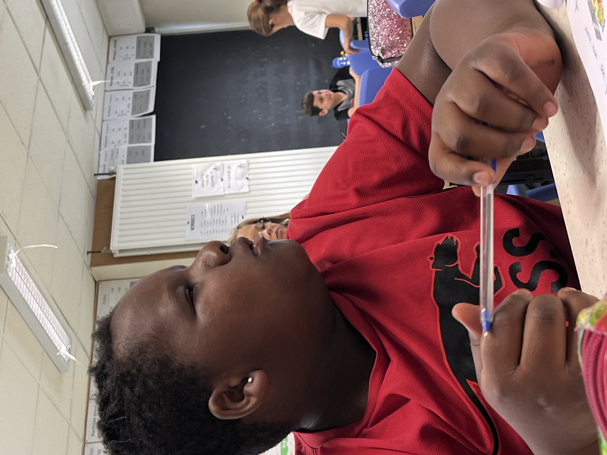 This image, captured in a classroom setting in La Fouillouse, France, features a person in the foreground, seen from a low, tilted angle. The main subject has dark skin and short, curly dark hair, wearing a red t-shirt adorned with a black, stylized animal logo on the chest, possibly a meerkat. They are holding a clear ballpoint pen in their right hand, with a focused gaze directed upwards and to the left. A small silver stud earring is visible in their left ear.
In the mid-ground, other individuals are present: a person with fair skin and glasses is partially visible to the right, and further back, a young girl in a white t-shirt stands near a blue chair, beside a pink, sparkly bag. Behind her, a boy in a black t-shirt is seated, looking towards the camera with a slight smile.
The setting is an educational environment with light-colored walls, white acoustic panels or radiators, and fluorescent lighting. A large black chalkboard is prominent in the background. Numerous papers are taped to the walls, some displaying text. Notably, one chart is titled "Présent - Prétérit" (Present - Preterite), indicating a French language lesson. Another paper asks "WHAT'S YOUR VALUE?". The main subject is at a light-colored desk, where a colorful paper is also visible. The scene suggests a classroom during a learning or study session, with students engaged in their work.
