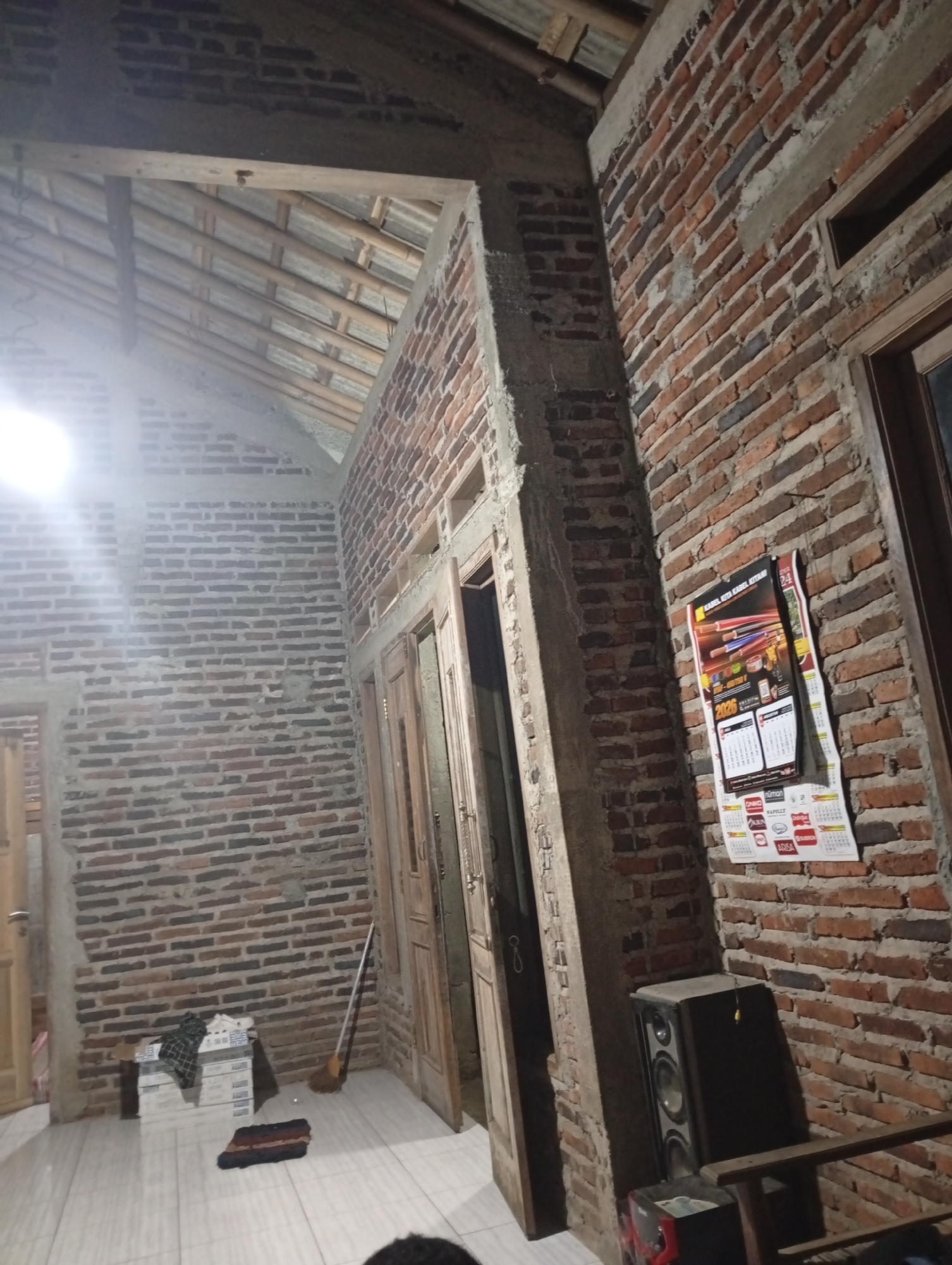 An indoor shot captures an unfinished brick room with wooden doors and a pitched roof. A large speaker sits on the floor next to a brick wall adorned with a calendar. A broom leans against the wall near a stack of boxes. The room appears to be under construction, with exposed brick and unfinished concrete elements. The lighting suggests it is either nighttime or the room is dimly lit.