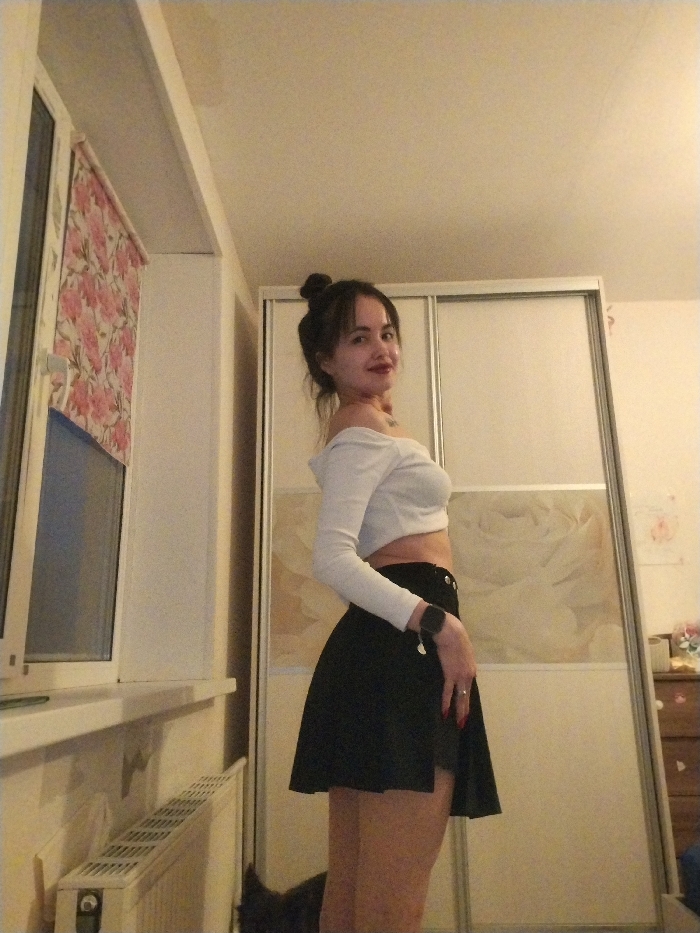 A young woman is captured in an indoor setting, likely a bedroom or living space, posing for the camera. She stands with her body slightly turned away, looking back over her left shoulder with a slight smile on her face, featuring red lipstick. Her dark hair is styled in a bun on top of her head, with some strands loose around her face.
She is wearing a white, off-the-shoulder, long-sleeved crop top that exposes her midriff, paired with a black A-line mini-skirt. On her left wrist, she wears a dark smartwatch, and a ring is visible on her left hand's ring finger. Her fingernails are painted red.
The background elements define the room. To the left of the woman, a white-framed window is visible, fitted with a roller blind adorned with a floral pattern of pink and red flowers. Below the window is a white windowsill, and beneath that, a white radiator with a grille pattern.
To the right, behind the woman, is a large wardrobe or closet with light-colored sliding doors that have a reflective or slightly translucent quality, showing faint, swirly patterns. Further to the right, a light-colored wall is visible, with some faint decorations or artwork near the top. Below this, a dark wooden dresser or chest of drawers can be seen.
The lighting suggests an indoor setting, possibly evening or night, with artificial illumination creating a warm atmosphere. The ceiling is a light, plain color, showing some minor textural variations. A small, dark object, possibly a pet or a piece of furniture, is partially visible at the very bottom center of the frame, near the floor.