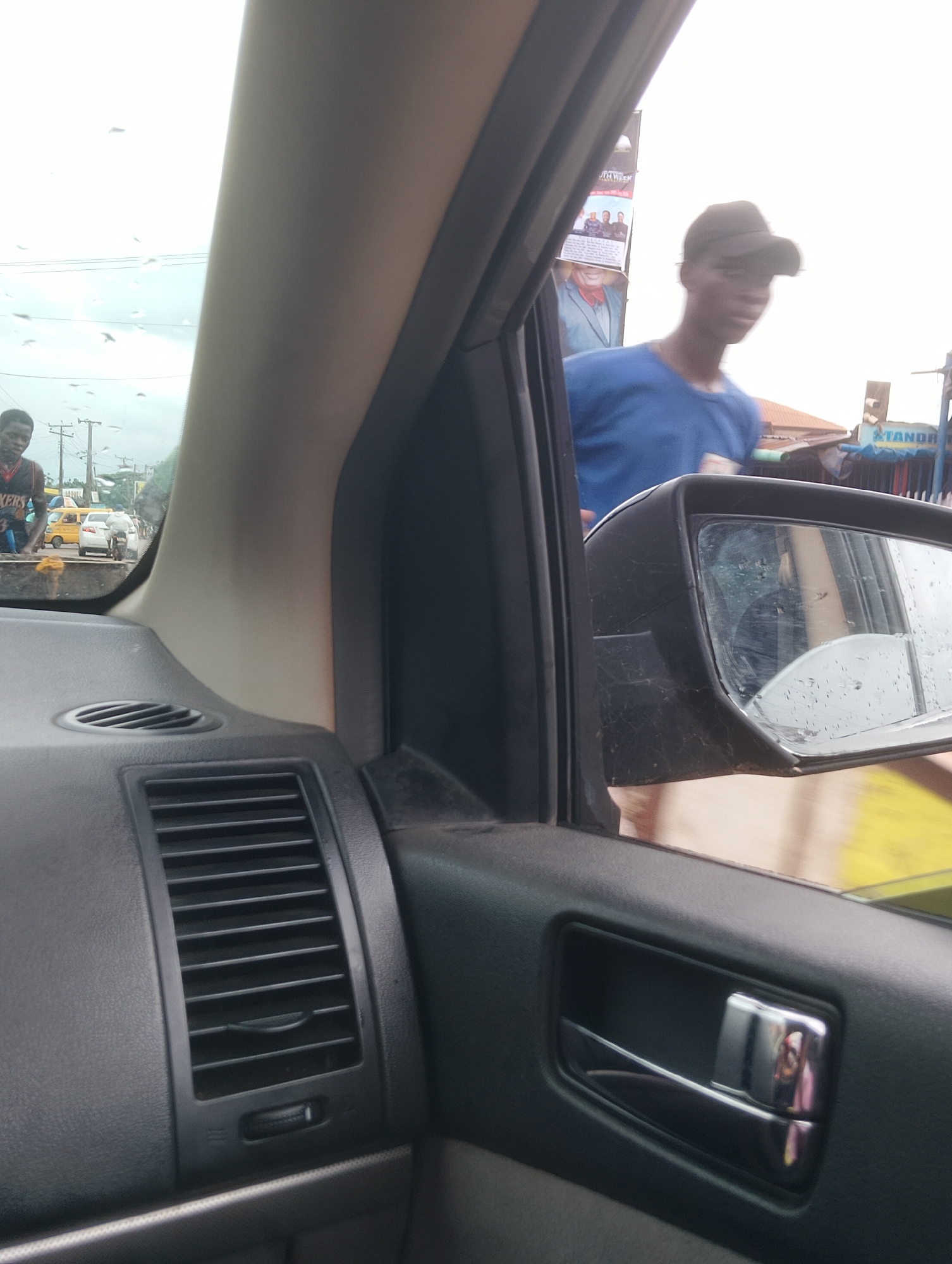The image, taken from inside a vehicle, offers a view of a street scene in Aketu-Oja, Nigeria. The foreground is dominated by the interior of the car, showing a black dashboard with air vents and a silver door handle. Through the passenger window, a young man in a blue shirt and a dark baseball cap is partially visible, looking away from the camera. Further in the background, another young man, wearing a sleeveless shirt, stands near a yellow vehicle and other pedestrians. The environment appears to be a busy street with signs and buildings lining the road. The sky is overcast, with some rain droplets visible on the windshield, suggesting recent or ongoing precipitation.