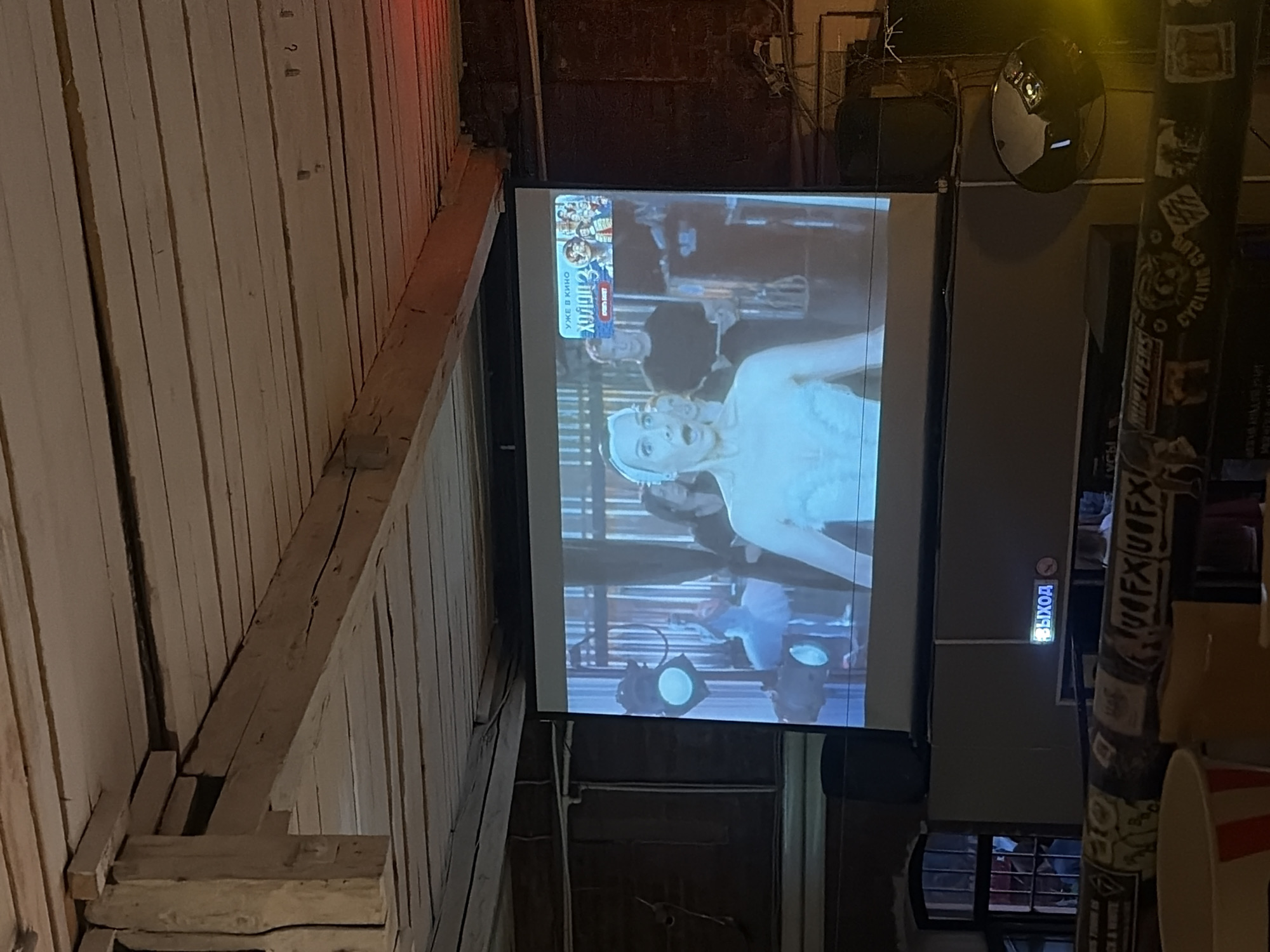 The image depicts an indoor scene where a projector screen displays a movie or show. The screen shows a woman in a white leotard and a ballet tutu, with her mouth open in surprise. Behind her, other people are visible, including a man in a black t-shirt. A small inset on the top right of the screen displays text in Russian, including "УЖЕ В КИНО" (Already in Cinema) and what appears to be a movie title. The projector screen is mounted against a dark wall, and the ceiling above is constructed of wooden beams and planks, suggesting an older building, possibly a bar or a repurposed industrial space. In the foreground, a dark railing is visible, adorned with various stickers, some of which are partially obscured. A small, illuminated sign with Russian text that reads "ВЫХОД" (Exit) is mounted on a wall below the projector screen. The overall ambiance suggests a casual setting where a film is being projected for an audience, likely in Saint Petersburg, Russia, given the Russian language on the screen and exit sign.