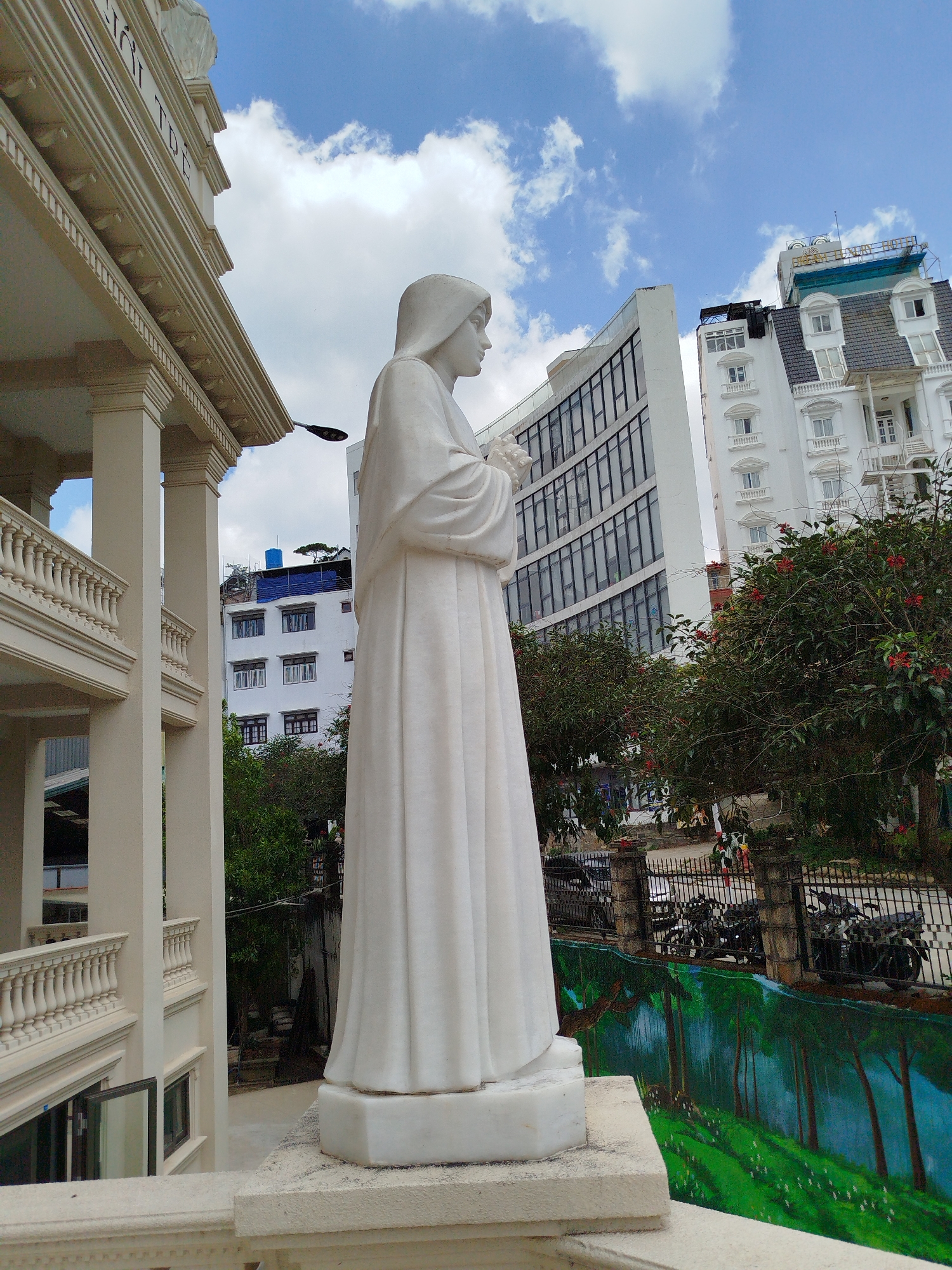 The image captures an outdoor scene in Da Lat, Vietnam, centered on a large white marble or stone statue of a woman. Dressed in a flowing, hooded robe, the statue holds a small bouquet of flowers with clasped hands, gazing towards the right. It stands on a raised platform, seemingly part of a balcony or terrace.
To the left, a grand, light-colored building with classical architectural features such as large columns, balustrades, and intricate cornices is prominently featured. Visible Vietnamese text on its upper facade clearly reads "NHÀ THỜ ĐÀLẠT" (Dalat Cathedral/Church).
The background reveals a bustling urban environment under a bright, cloudy daytime sky. Directly behind the statue is a modern building with a curved glass facade and dark-framed windows. Further back and to the right, a multi-story white building with an older architectural style, featuring numerous balconies and a dark, possibly mansard, roof, adds to the diverse urban landscape.
Lush green trees with red blossoms are scattered between the buildings and in the mid-ground. Below them, a dark metal fence lines what appears to be a street or parking area, where several motorcycles are visible. A distinctive mural adorns a wall below the fence, depicting a serene green forest or valley with tall trees and white flowers.
The photograph skillfully contrasts the classic beauty and spiritual symbolism of the statue and its accompanying architectural structure with the dynamic blend of modern development and natural elements characteristic of Da Lat.