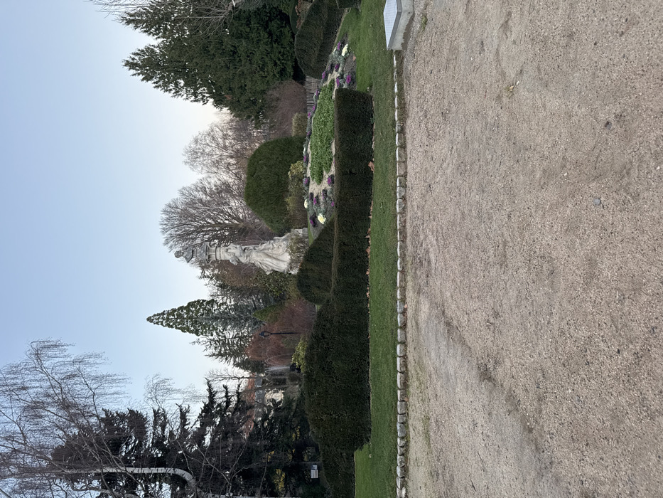 A park-like setting with a stone path along the right. Well-manicured hedges and shrubs line the path. A statue stands among bare trees in the center-left. Evergreen trees are visible in the background. The sky is light blue.